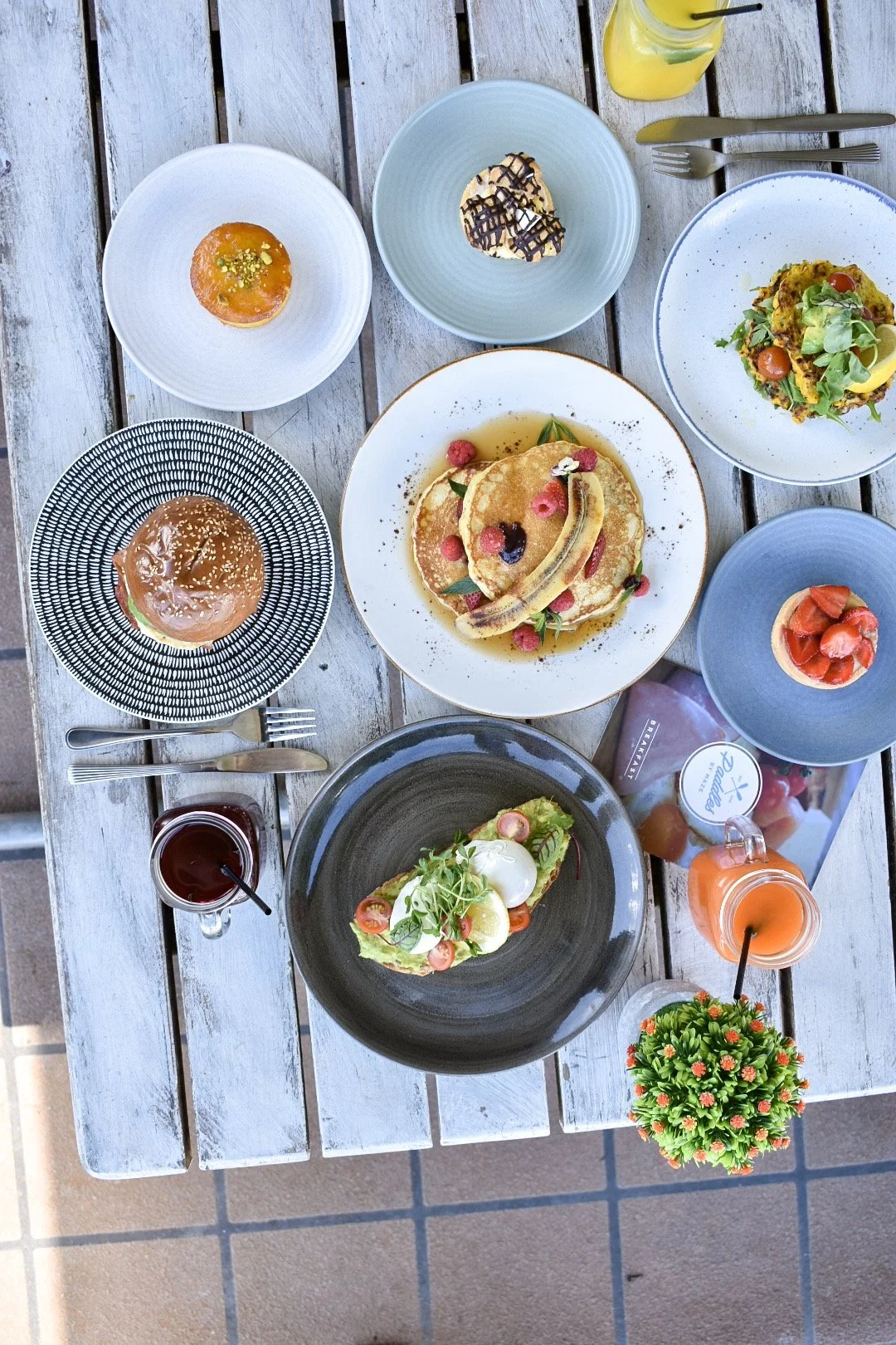 Overhead view of a brunch spread on a rustic wooden table, featuring plates of pancakes with berries and whipped cream, salads, pastries, and various drinks.