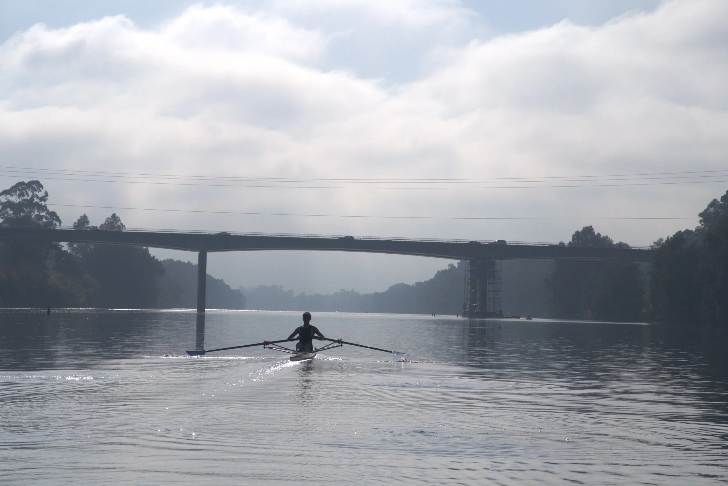 Person kayaking on a calm river or lake with a bridge and trees in the background, under a cloudy sky.