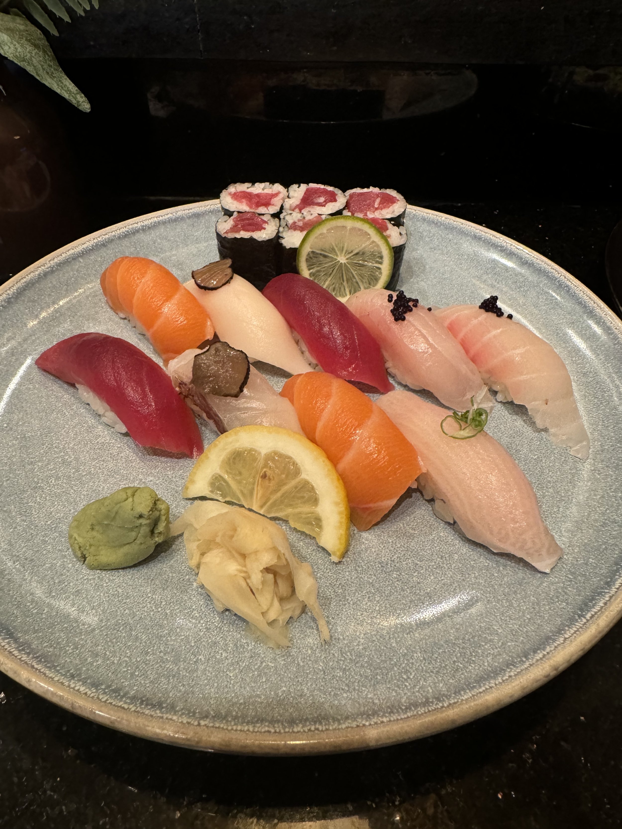 A plate of assorted sushi including various types of sashimi, sushi rolls, lemon slices, wasabi, and ginger, arranged on a ceramic plate.