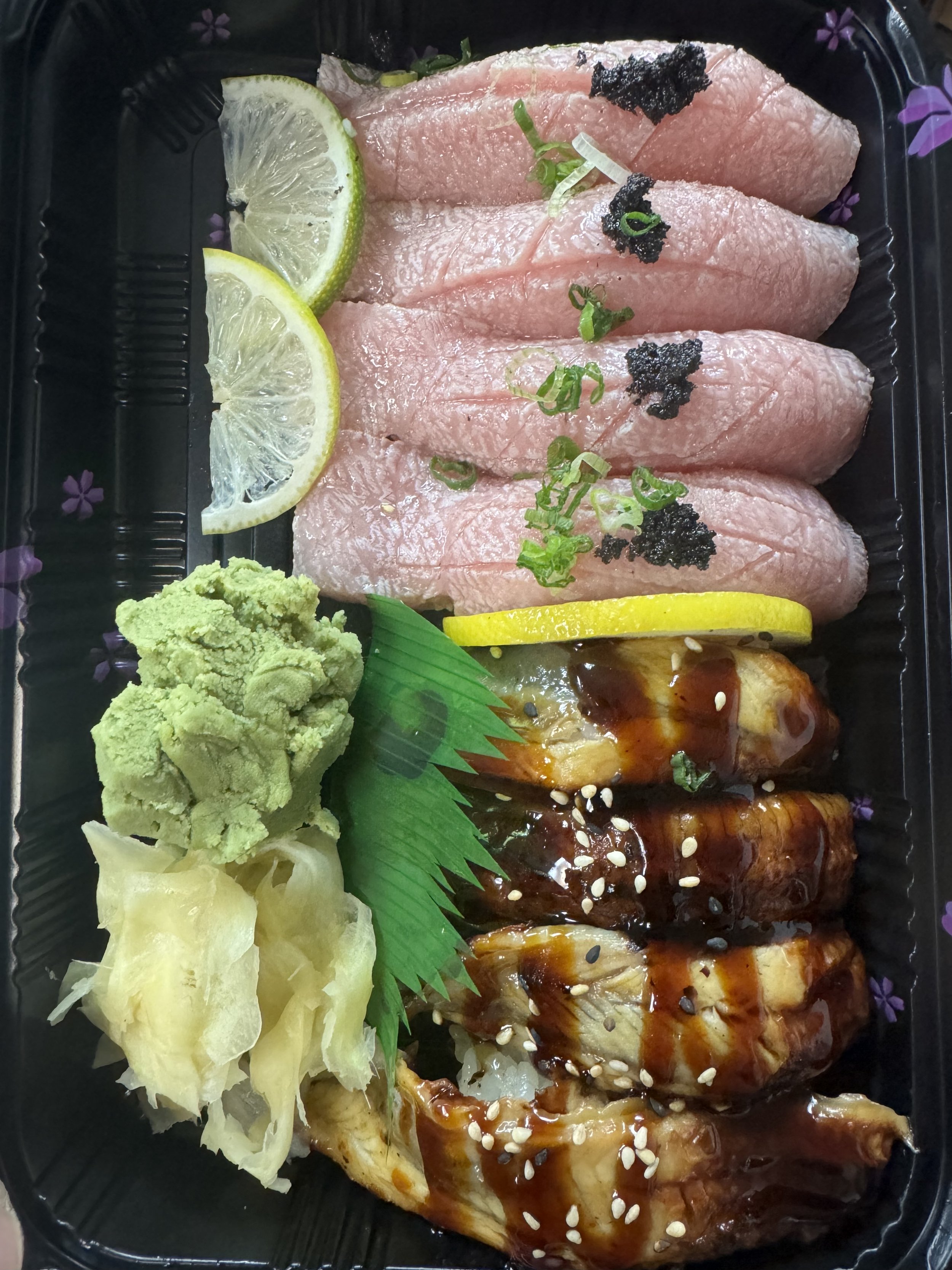 Sushi meal with slices of raw fish, lemon wedges, wasabi, pickled ginger, and a sushi roll with eel sauce in a black takeout container.