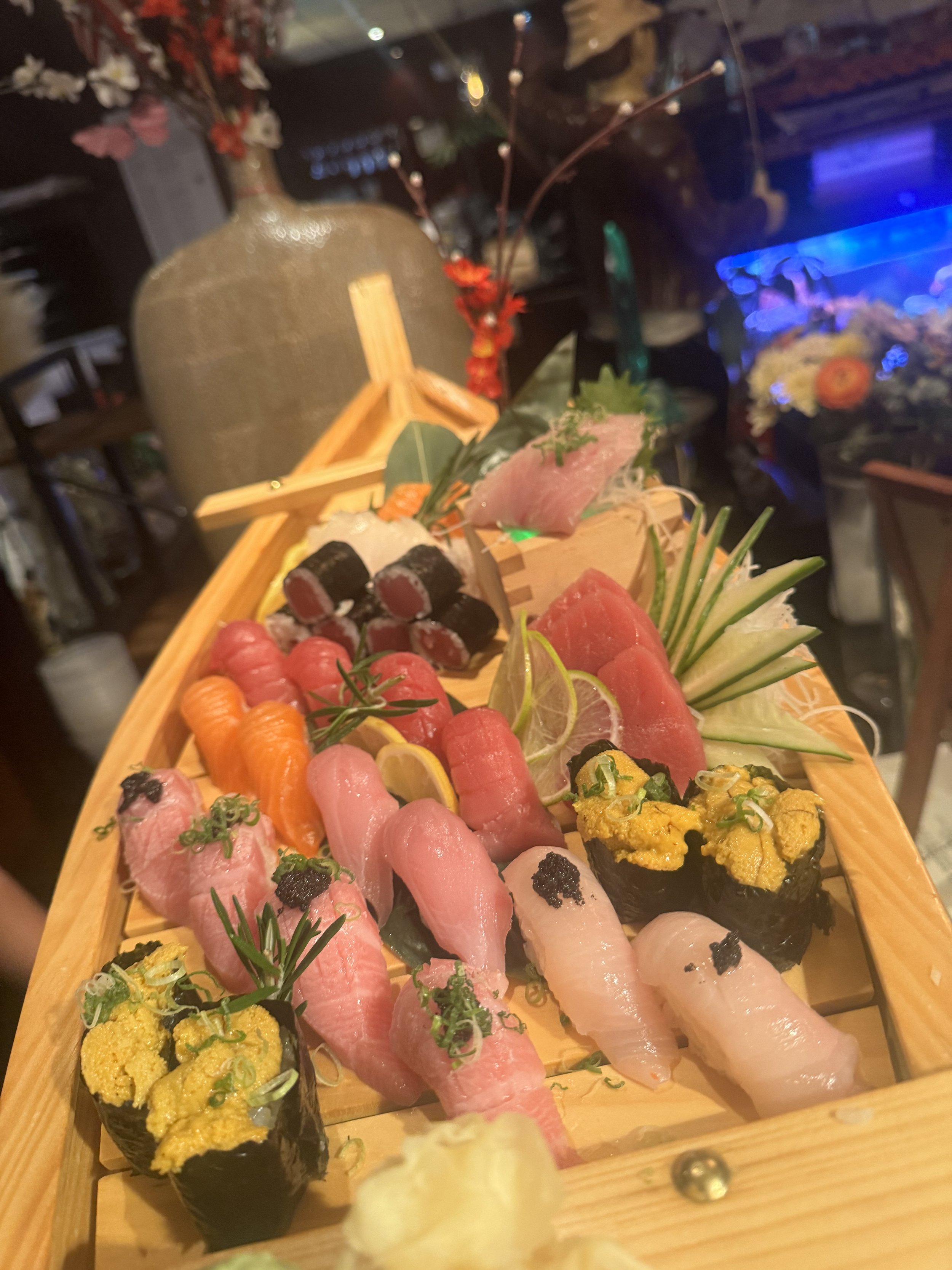 A wooden tray of assorted sushi and sashimi, including salmon, tuna, and other seafood, garnished with lemon slices, green leaves, and wasabi, set on a restaurant table.