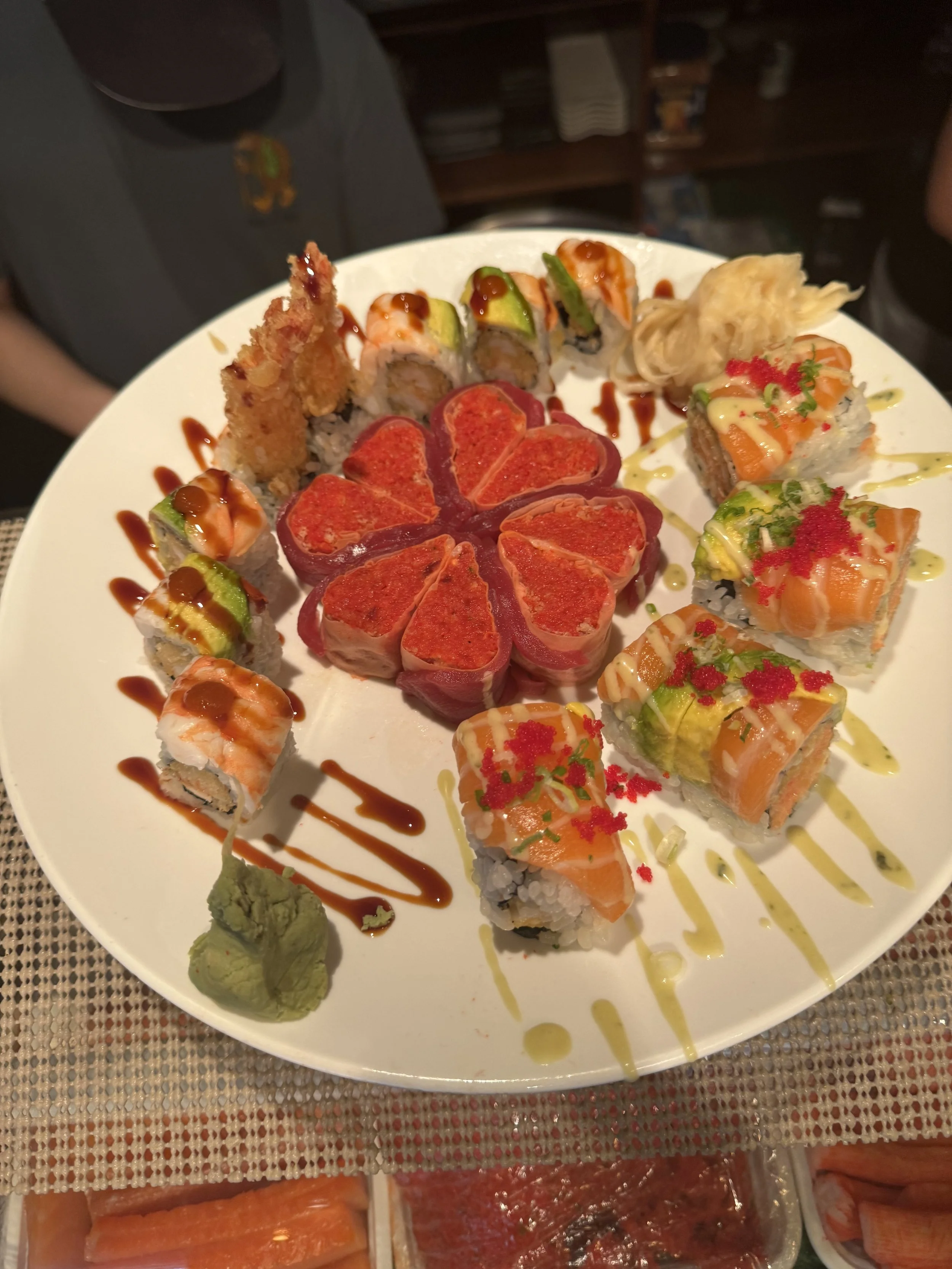 A white plate with assorted sushi rolls, including nigiri, topped with fish, avocado, and sauces, garnished with wasabi and ginger, on a woven placemat.