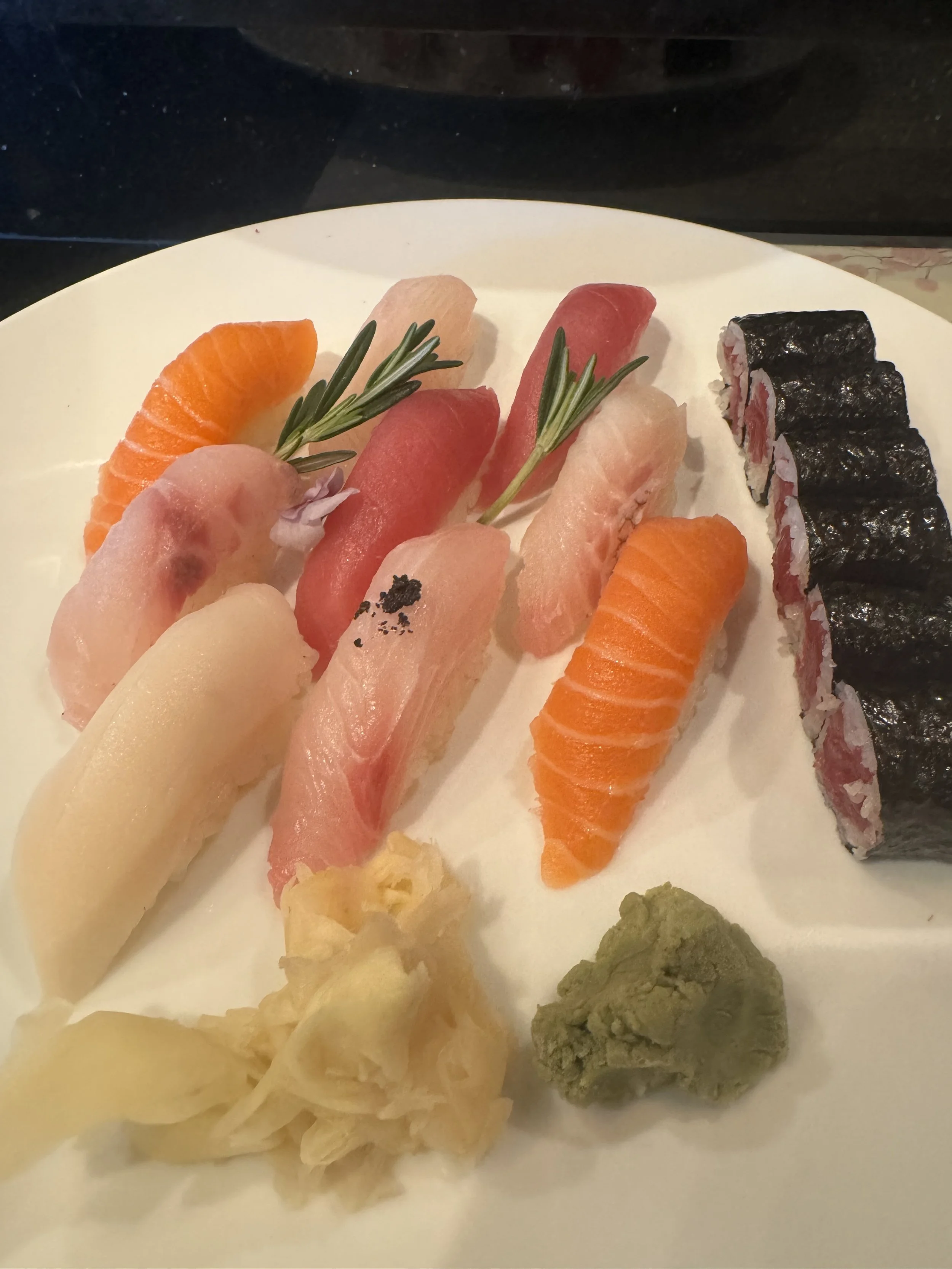 Plate of assorted sushi including salmon, tuna, and cucumber rolls, garnished with sprigs of rosemary, wasabi, and pickled ginger.