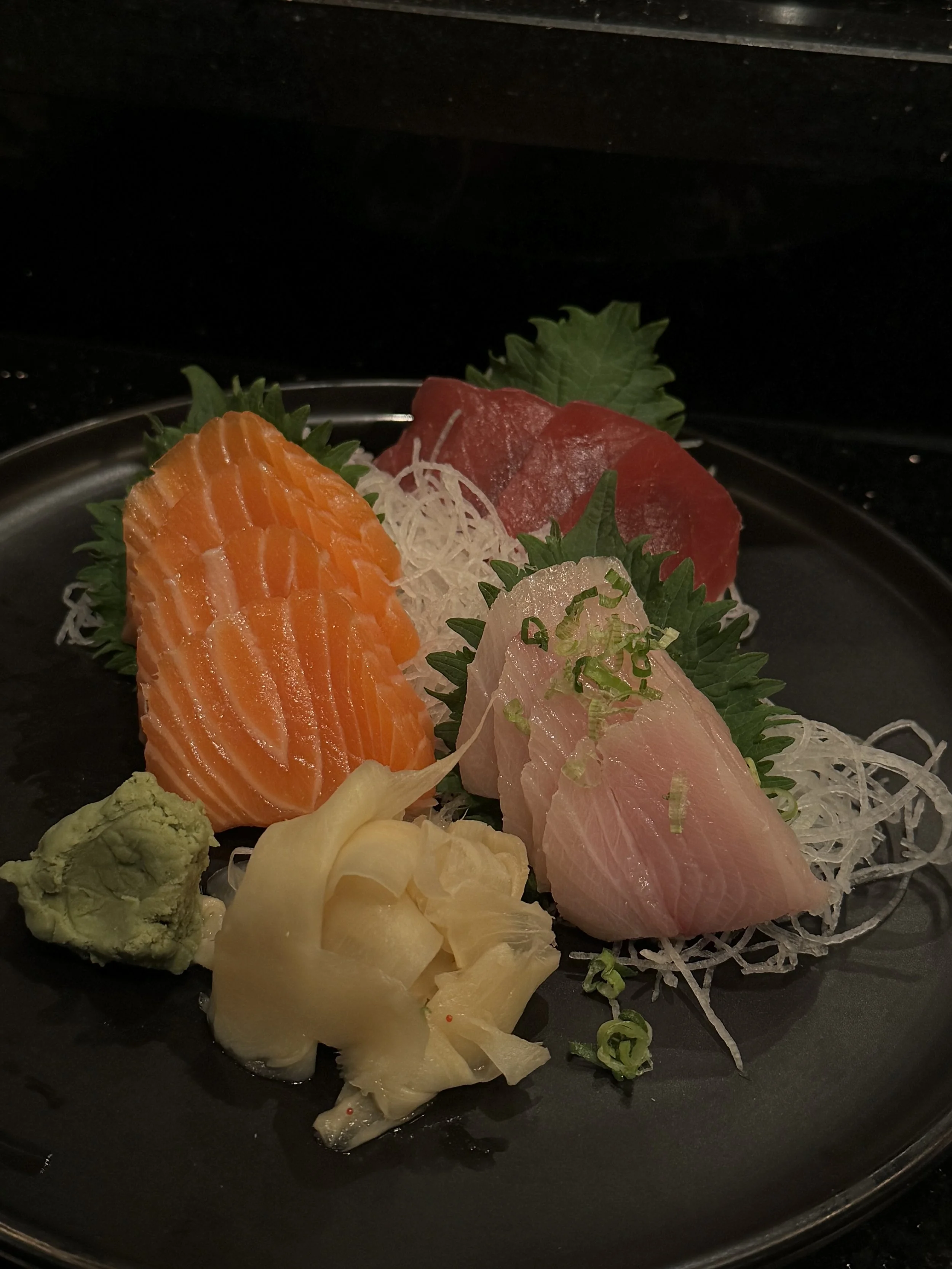 Assorted slices of sashimi, including salmon, tuna, and white fish, garnished with shredded daikon, green shiso leaves, wasabi, and pickled ginger, on a black plate.