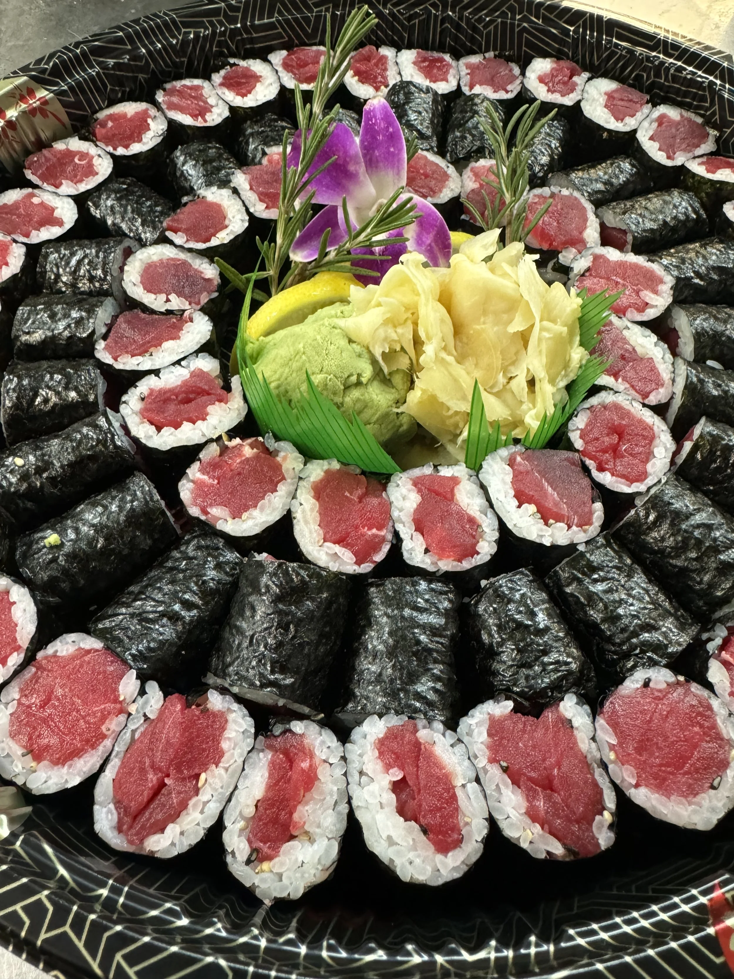 A tray of sushi rolls made with tuna, wrapped in nori seaweed, and accompanied by a dollop of wasabi, some pickled ginger, and a purple orchid flower for decoration.