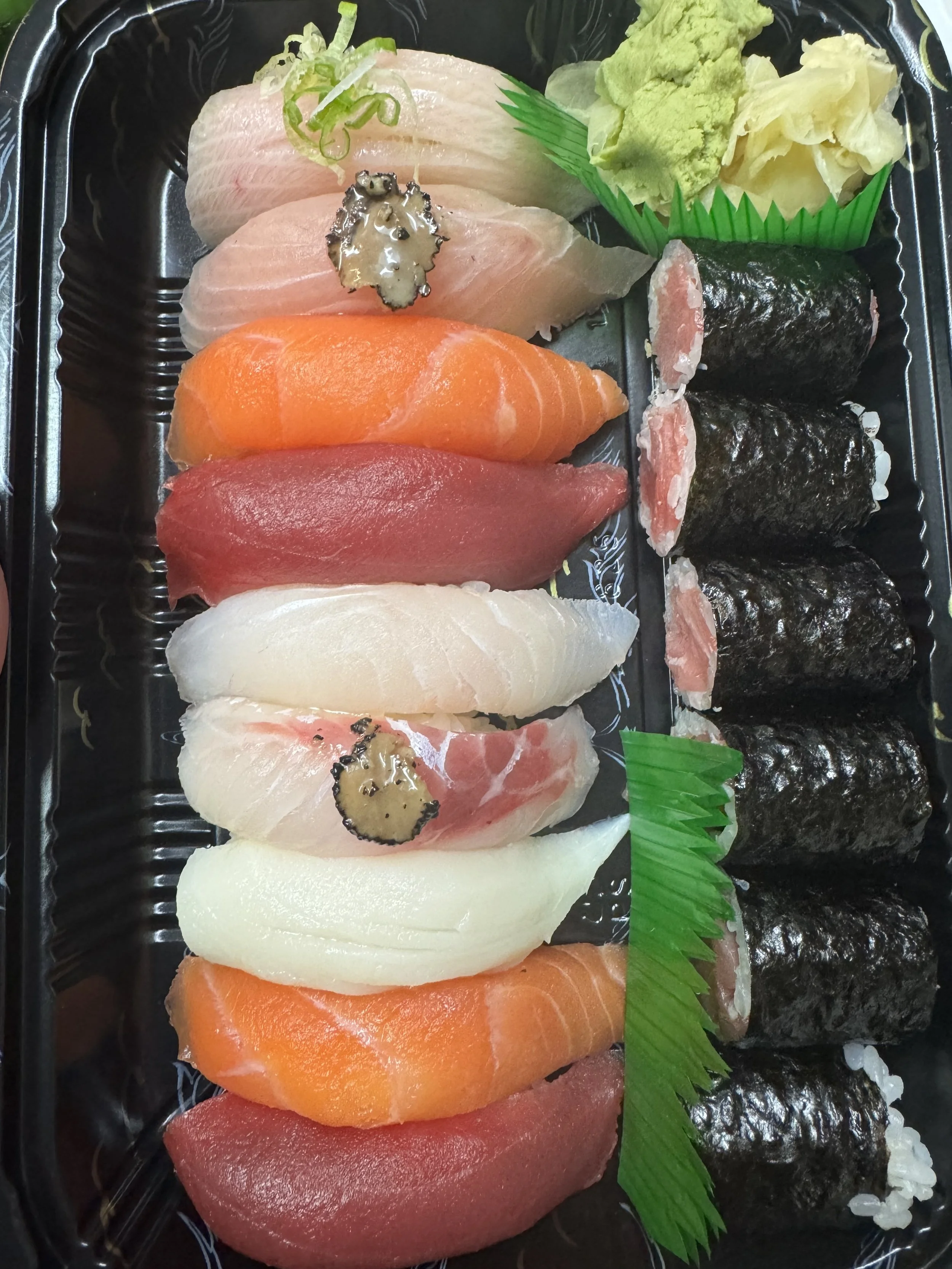 Assorted nigiri sushi with slices of raw fish including salmon, tuna, white fish, and yellowtail, garnished with wasabi, sliced ginger, and green plastic divider, served on a black tray.