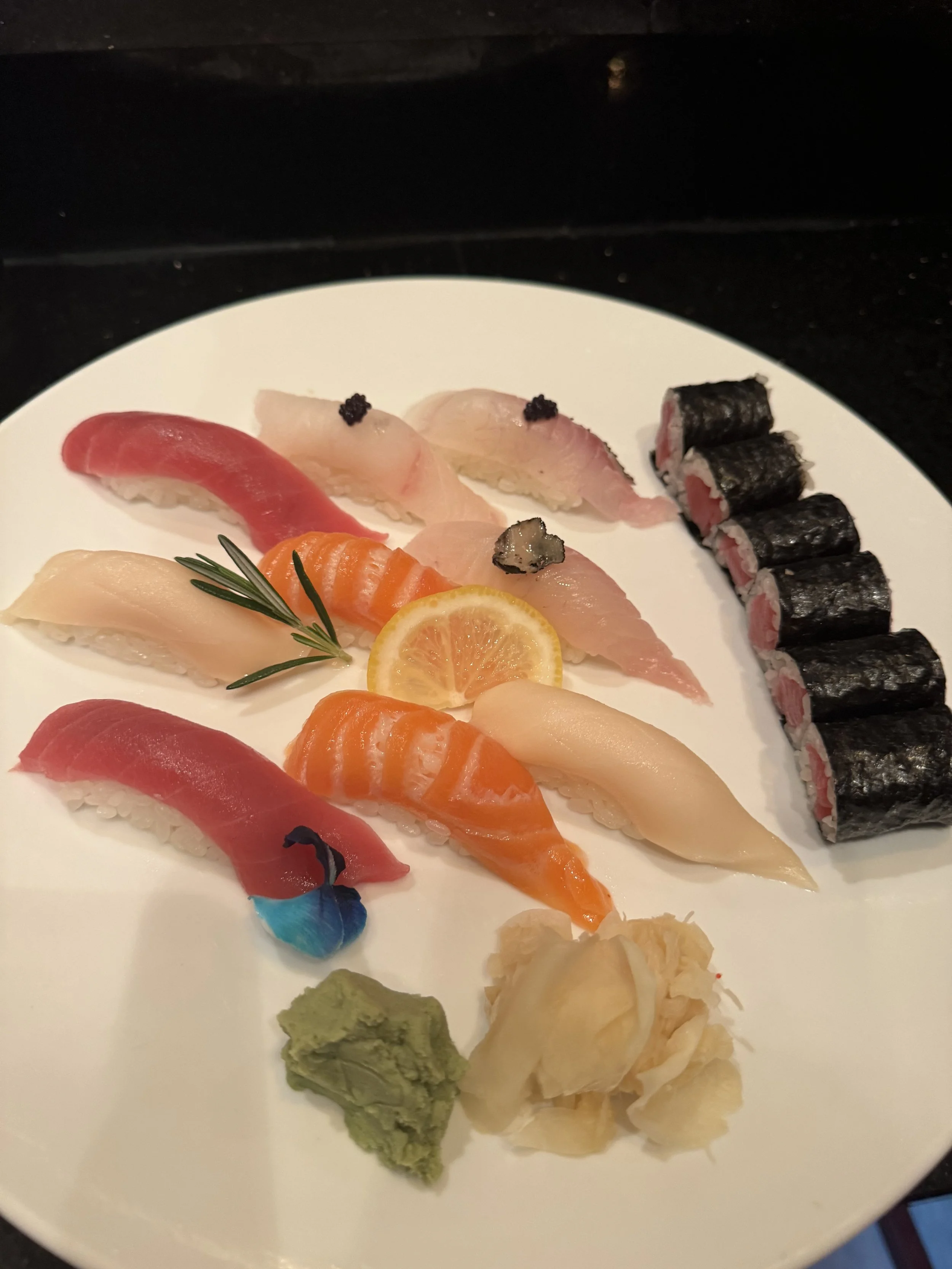 Assorted sushi on a white plate including sashimi with lemon slices, wasabi, pickled ginger, and seaweed-wrapped rolls.