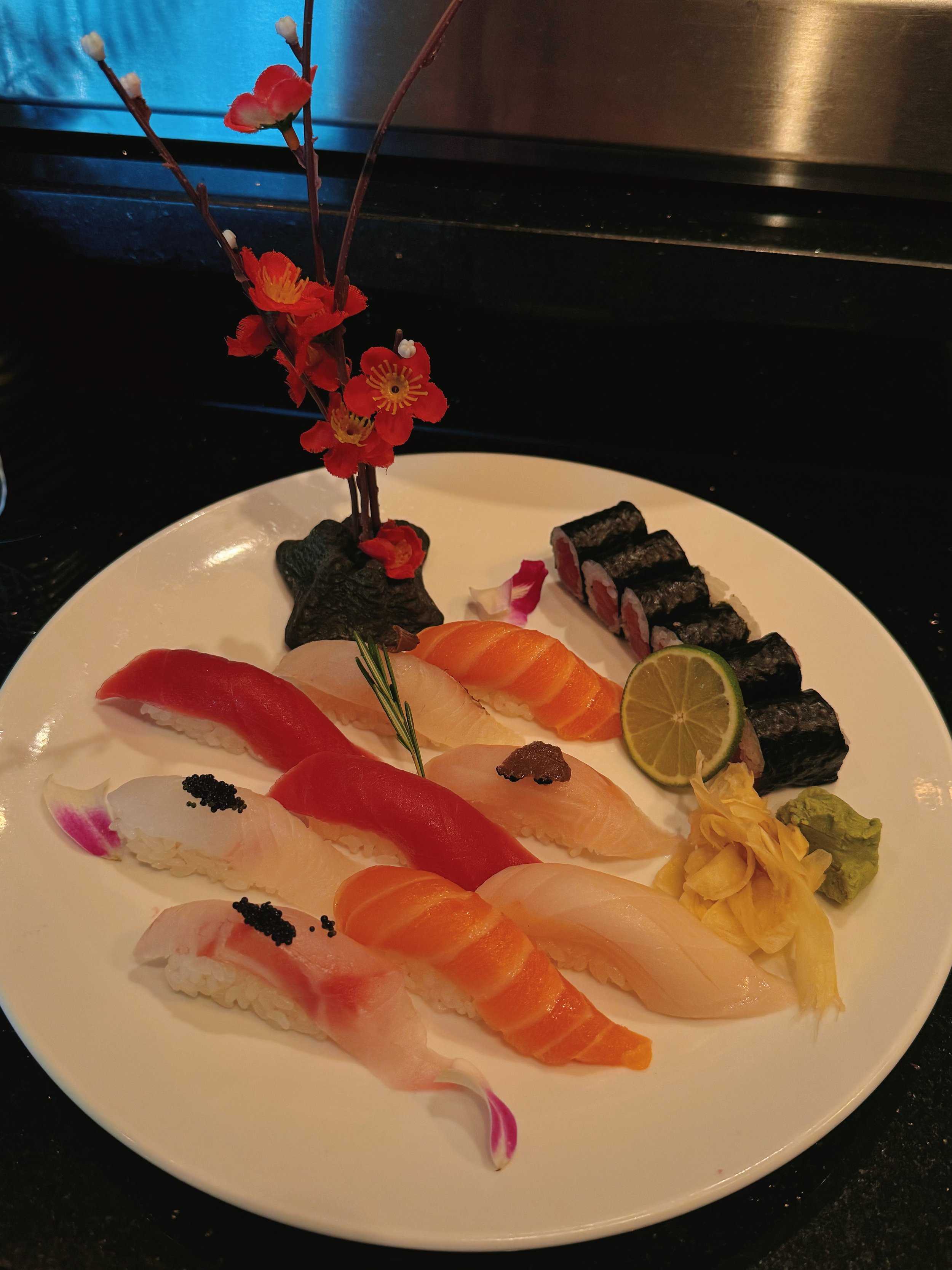 A white plate with assorted sashimi, including slices of salmon, tuna, and white fish, garnished with lime, wasabi, and yellow flower, and a decorative flower arrangement.