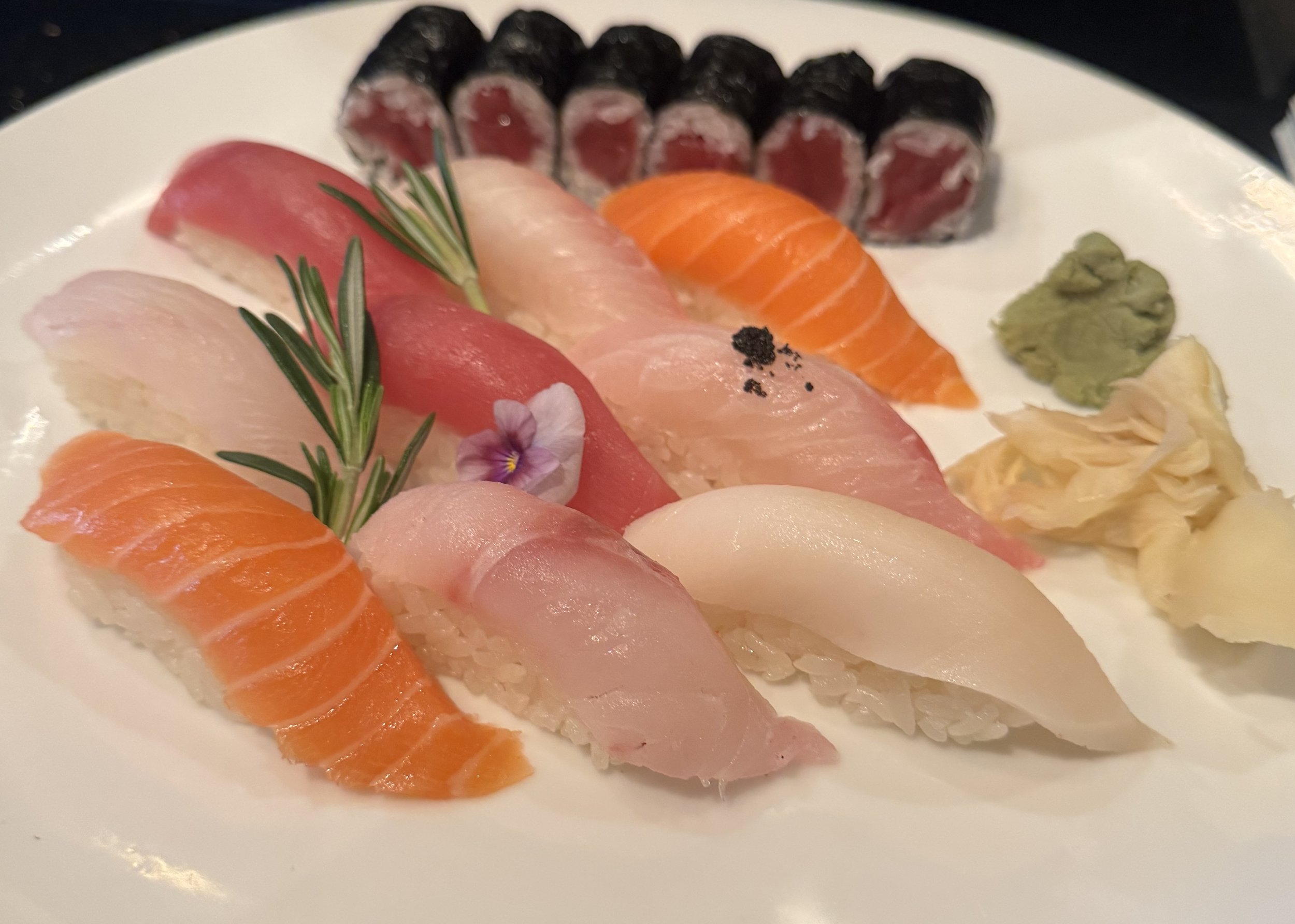 A white plate with assorted sushi including salmon, tuna, and white fish, garnished with a sprig of rosemary, a small amount of wasabi, and pickled ginger.