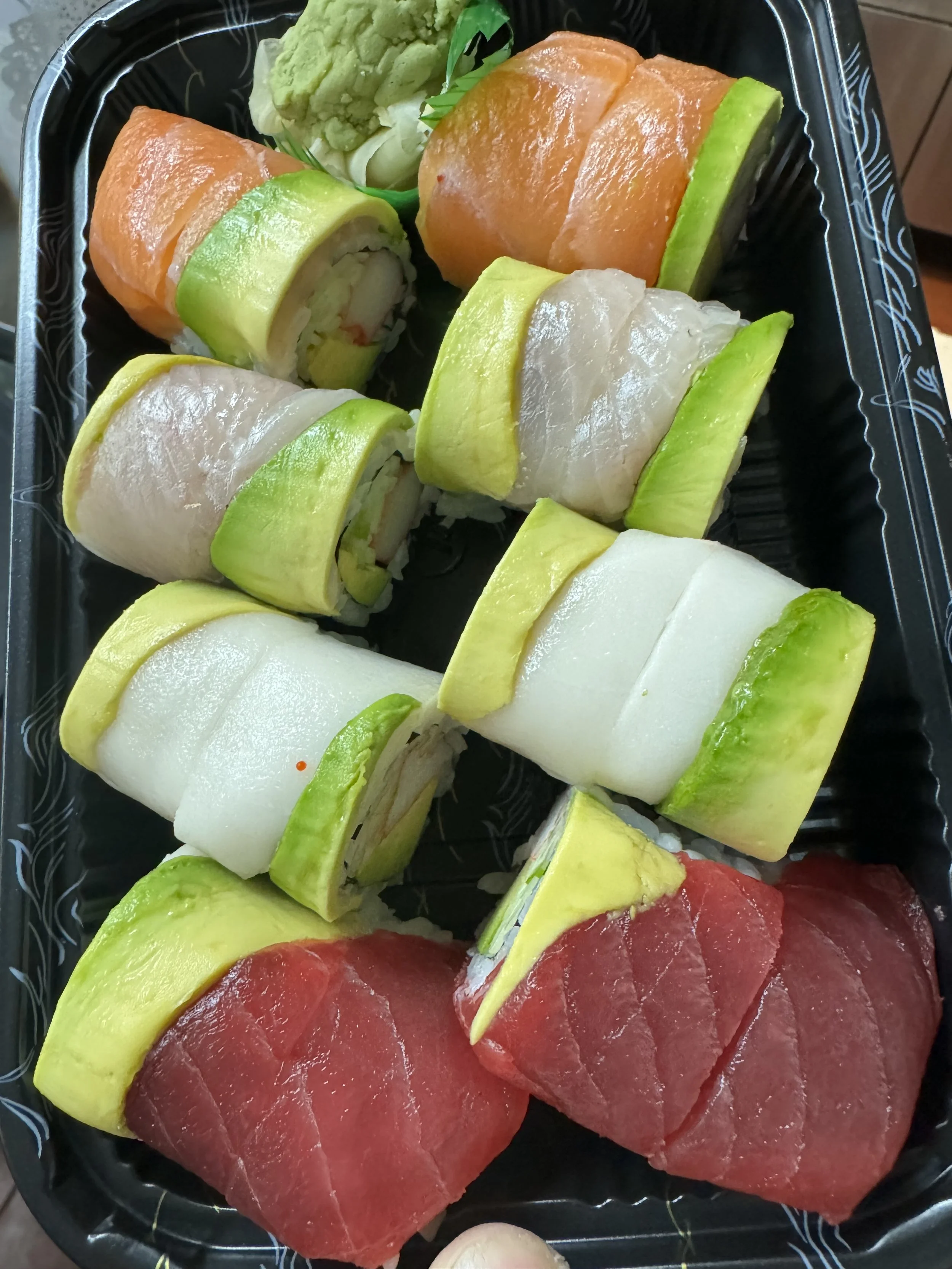 Assorted sushi rolls with slices of avocado, cucumber, and raw fish on a black plastic tray.