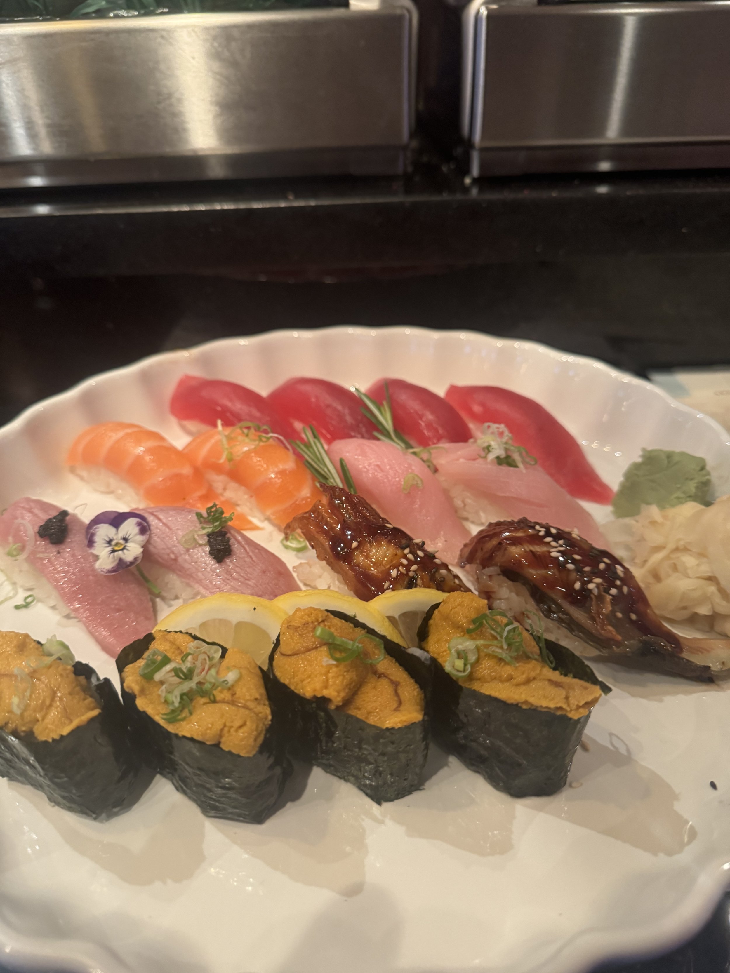 A white platter of assorted sushi including nigiri with salmon, tuna, and other fish, as well as sushi rolls topped with sea urchin, all garnished with herbs and flowers.