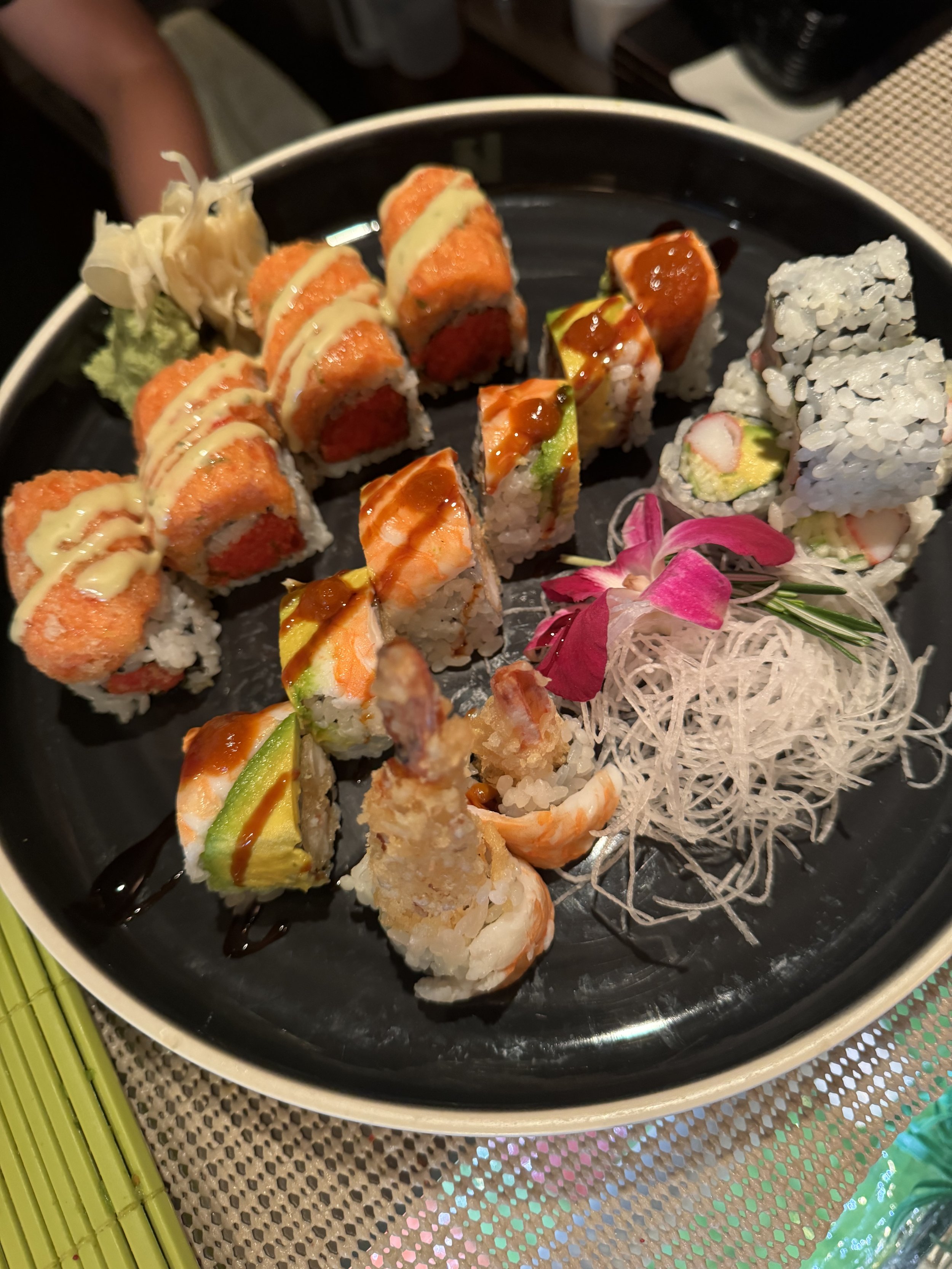 A variety of sushi rolls on a black plate, garnished with pink flower petals and shredded daikon, with wasabi and pickled ginger on the side.