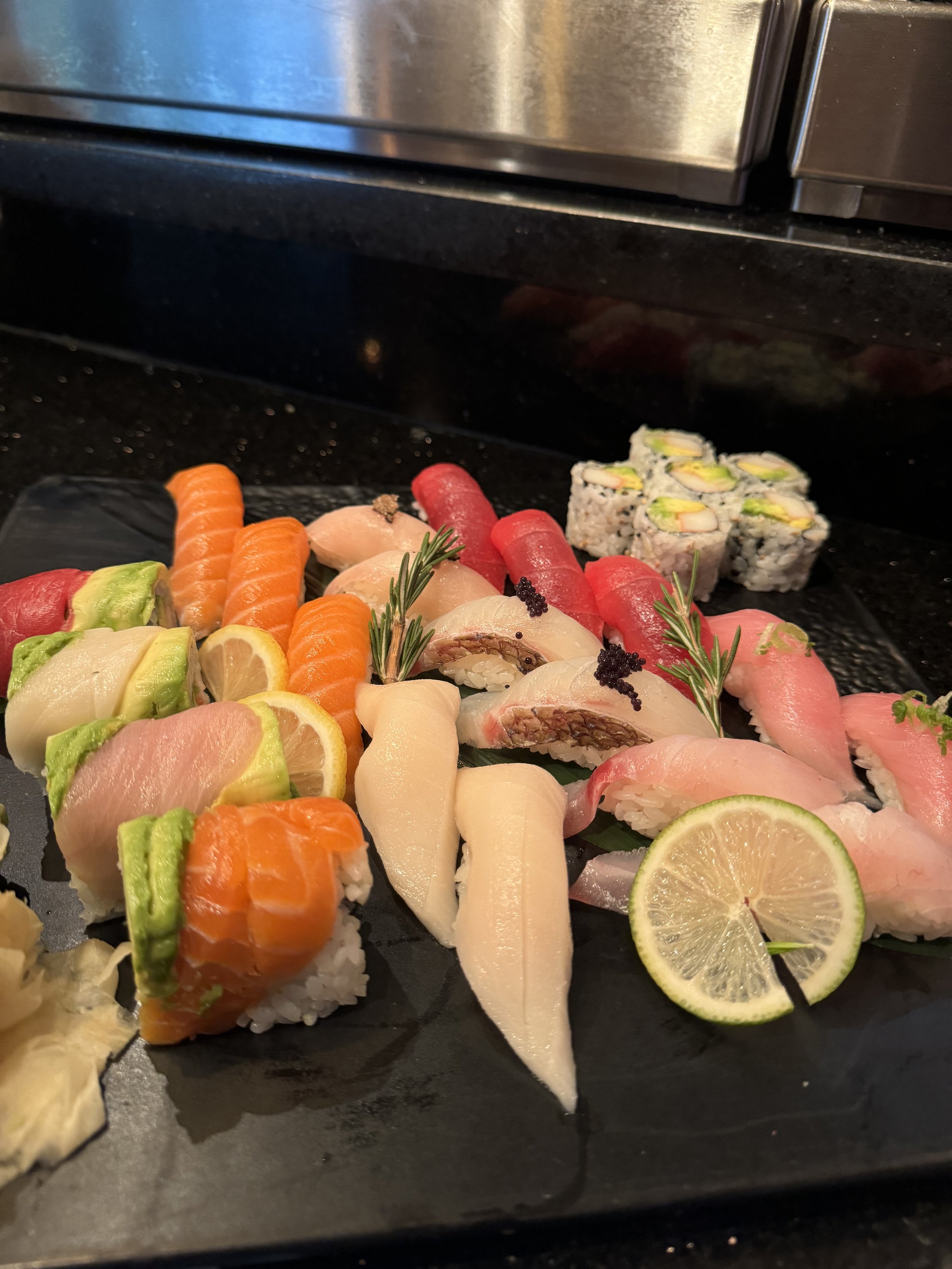 Assorted sushi rolls and sashimi with citrus slices, garnished with herbs on a black platter.