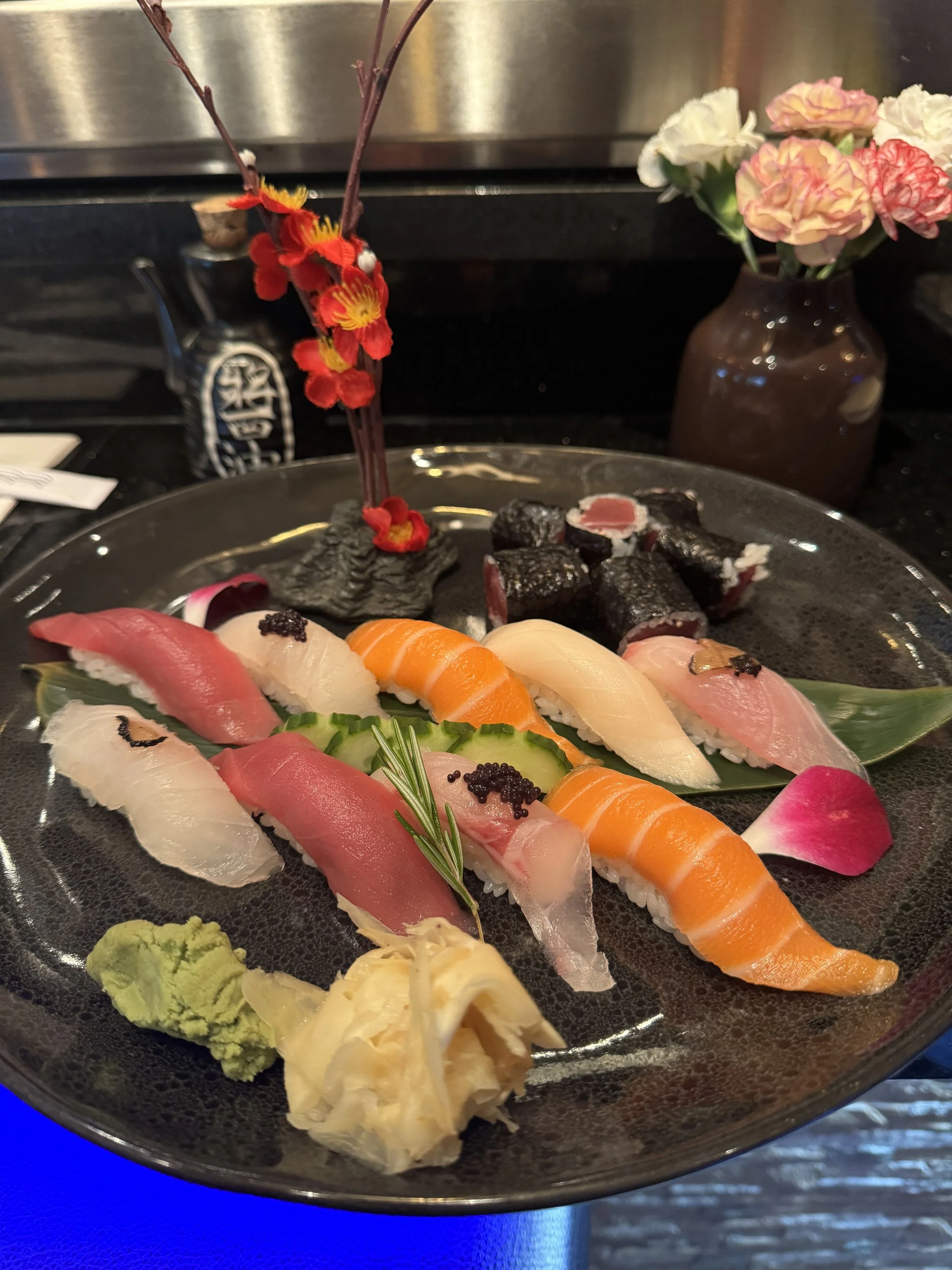 Assorted sushi pieces including tuna, salmon, and white fish with garnishes of wasabi, pickled ginger, and a sprig of rosemary on a black plate.