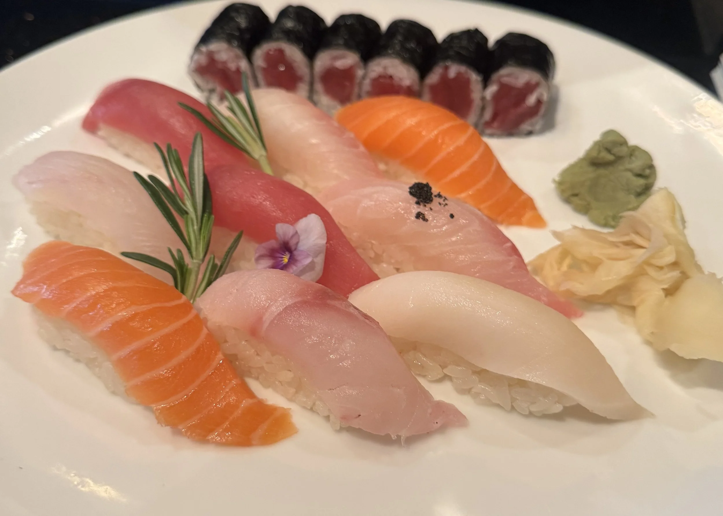 Assorted sushi slices on a white plate, garnished with green herbs, a flower, wasabi, and pickled ginger.
