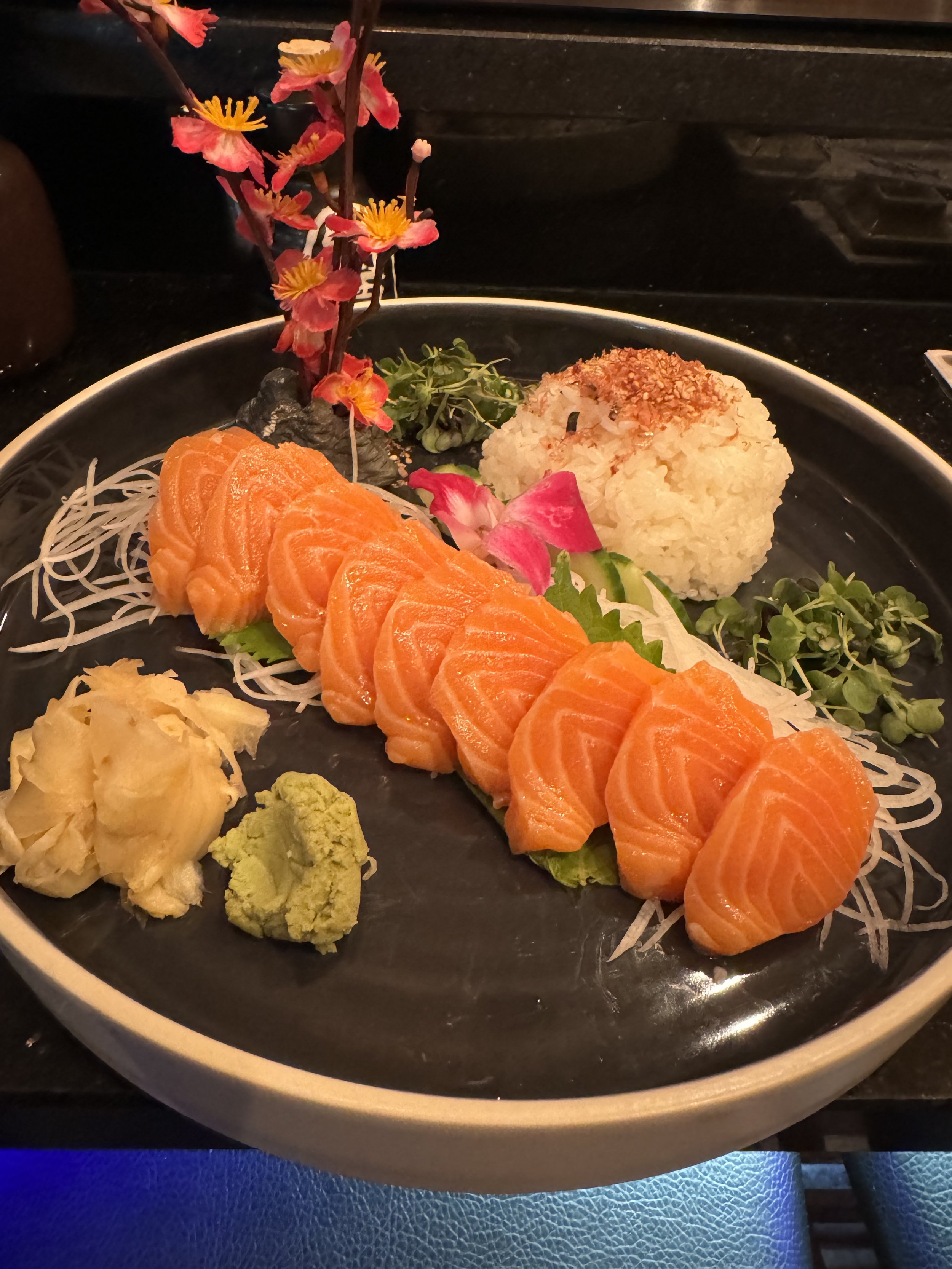 A plate of sashimi featuring sliced salmon, wasabi, pickled ginger, and a rice ball garnished with chopped nuts, alongside decorative flowers, microgreens, and shredded daikon radish.
