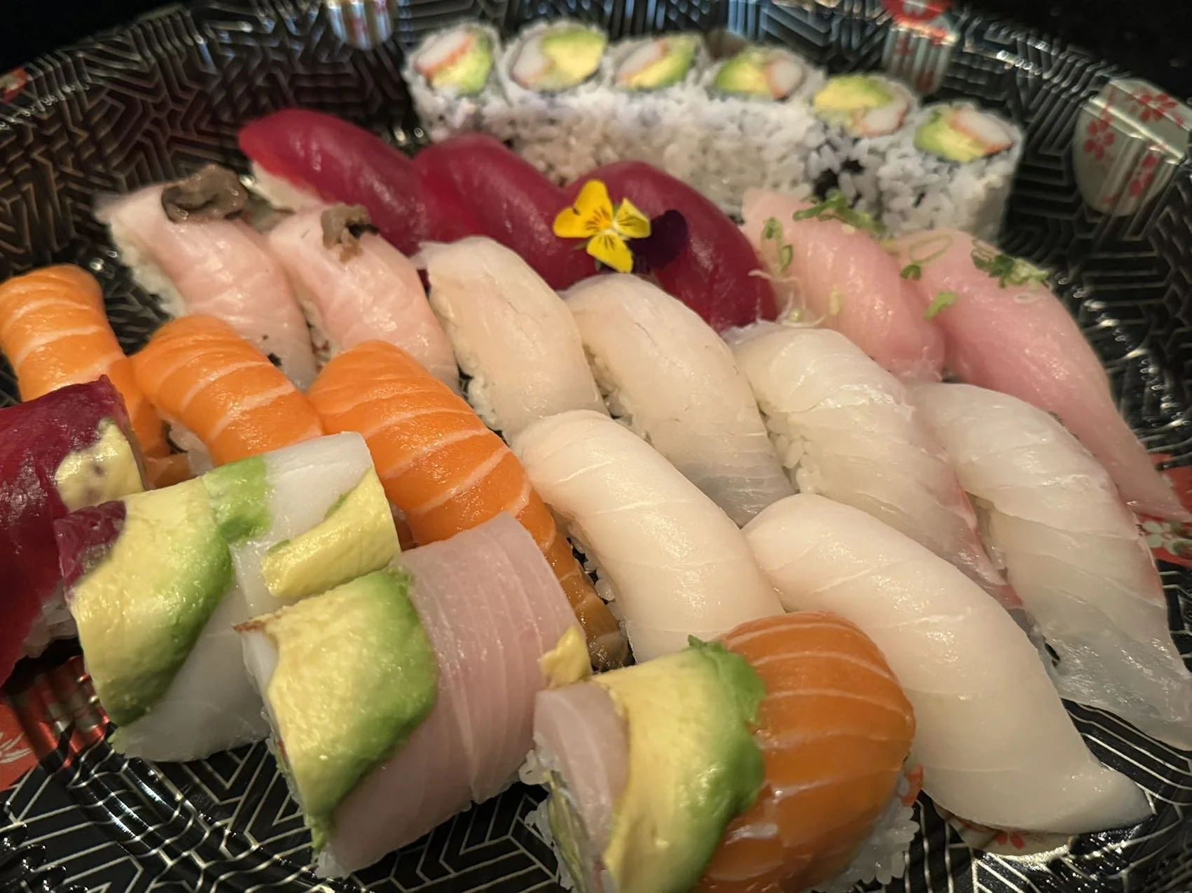 Assorted sushi including tuna, salmon, yellowtail, and cucumber rolls on a black patterned tray.