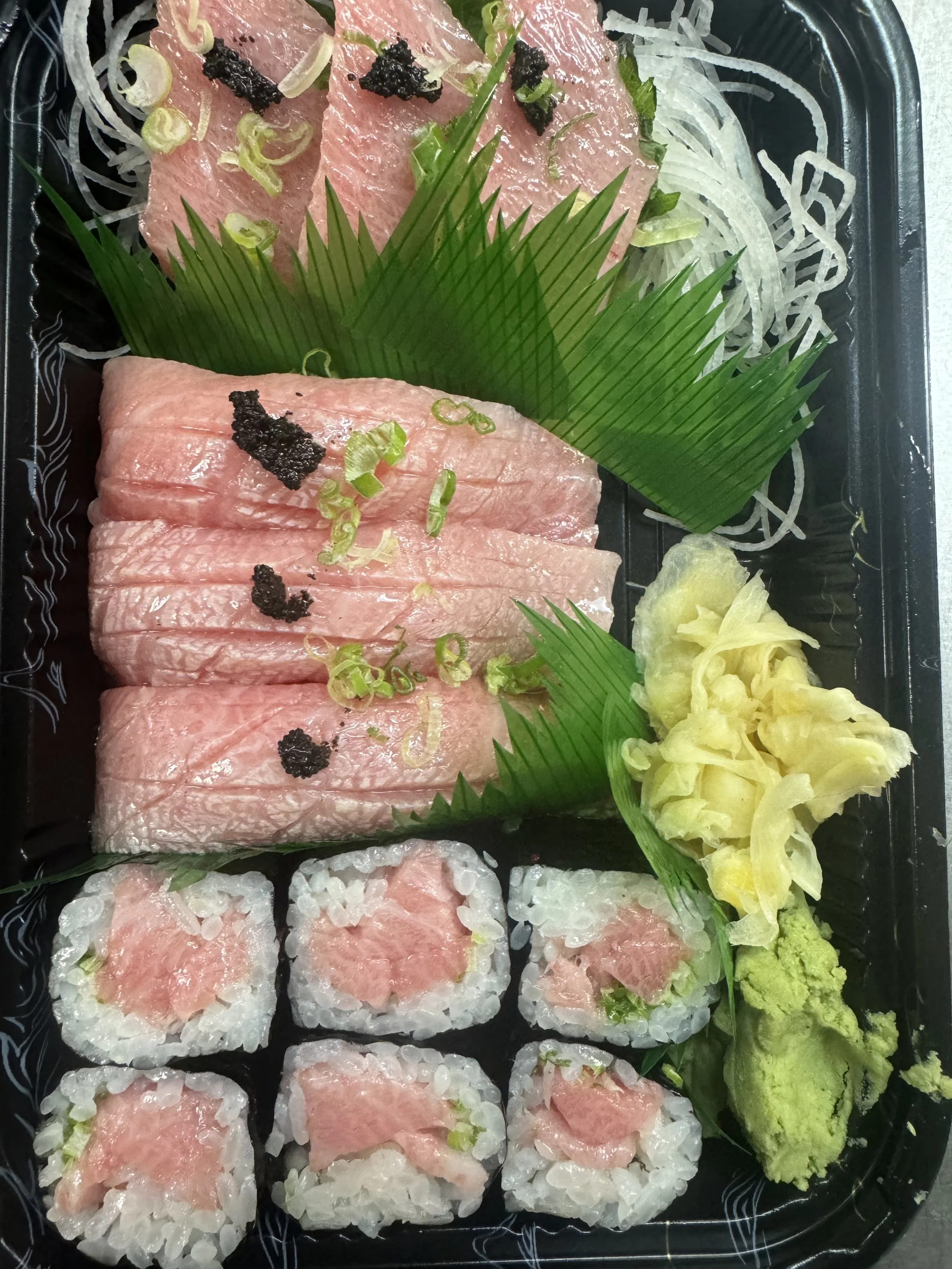 Sushi platter featuring slices of raw tuna sashimi garnished with chopped green onions and black garnish, green decorative leaves, shredded daikon radish, wasabi, and pickled ginger.