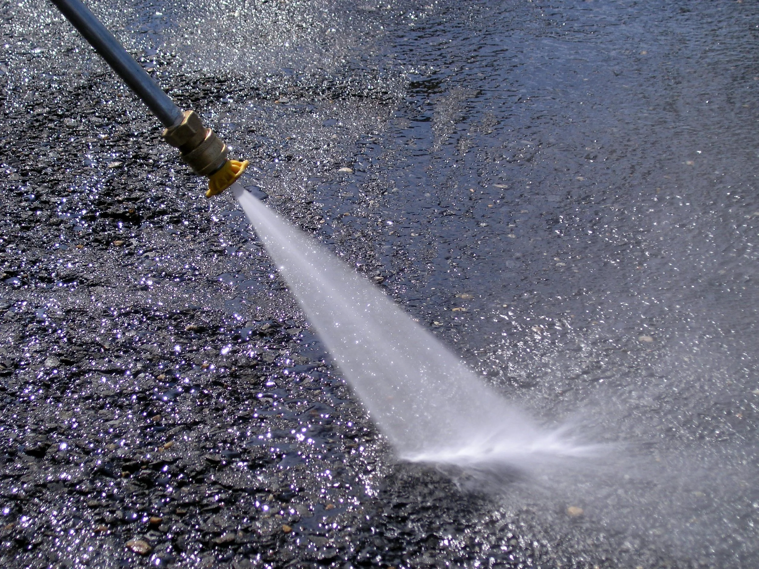 A pressure washer spraying water on an asphalt surface, causing water and debris to spray outward, with highlights reflecting sunlight.