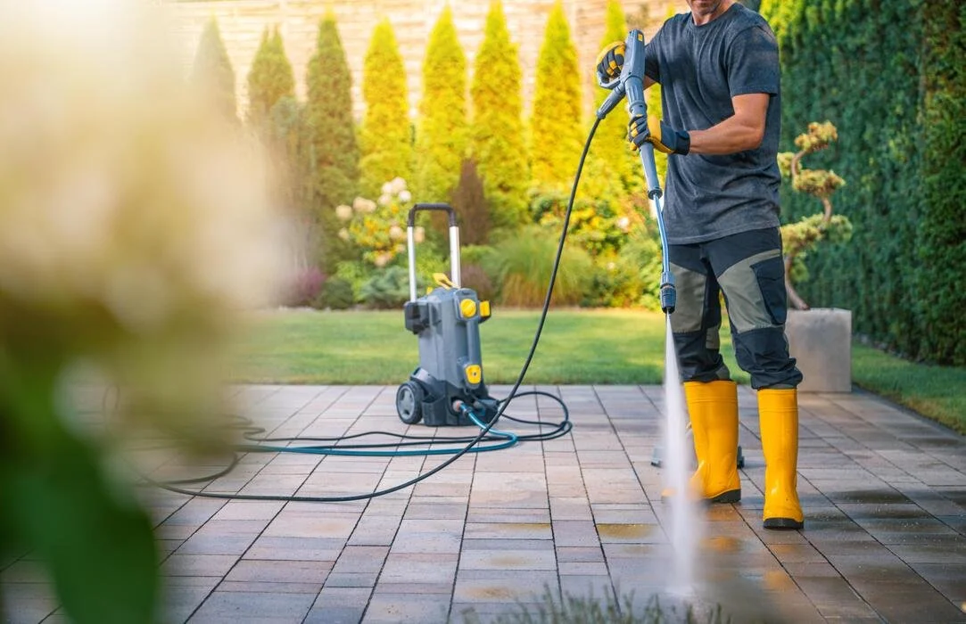 Person using a power washer to clean the patio in a backyard with green shrubs and trees.