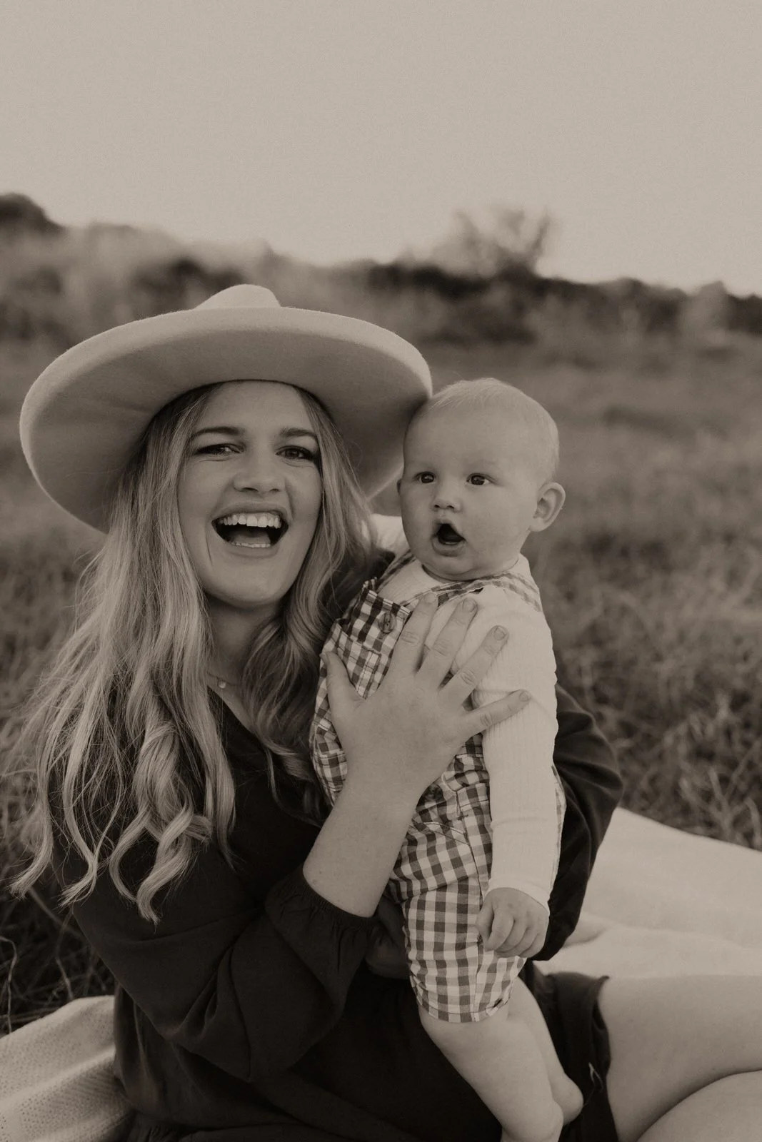 A woman wearing a large hat holding a young child in an open field, both looking off to the side with the woman smiling and the child with a surprised expression.