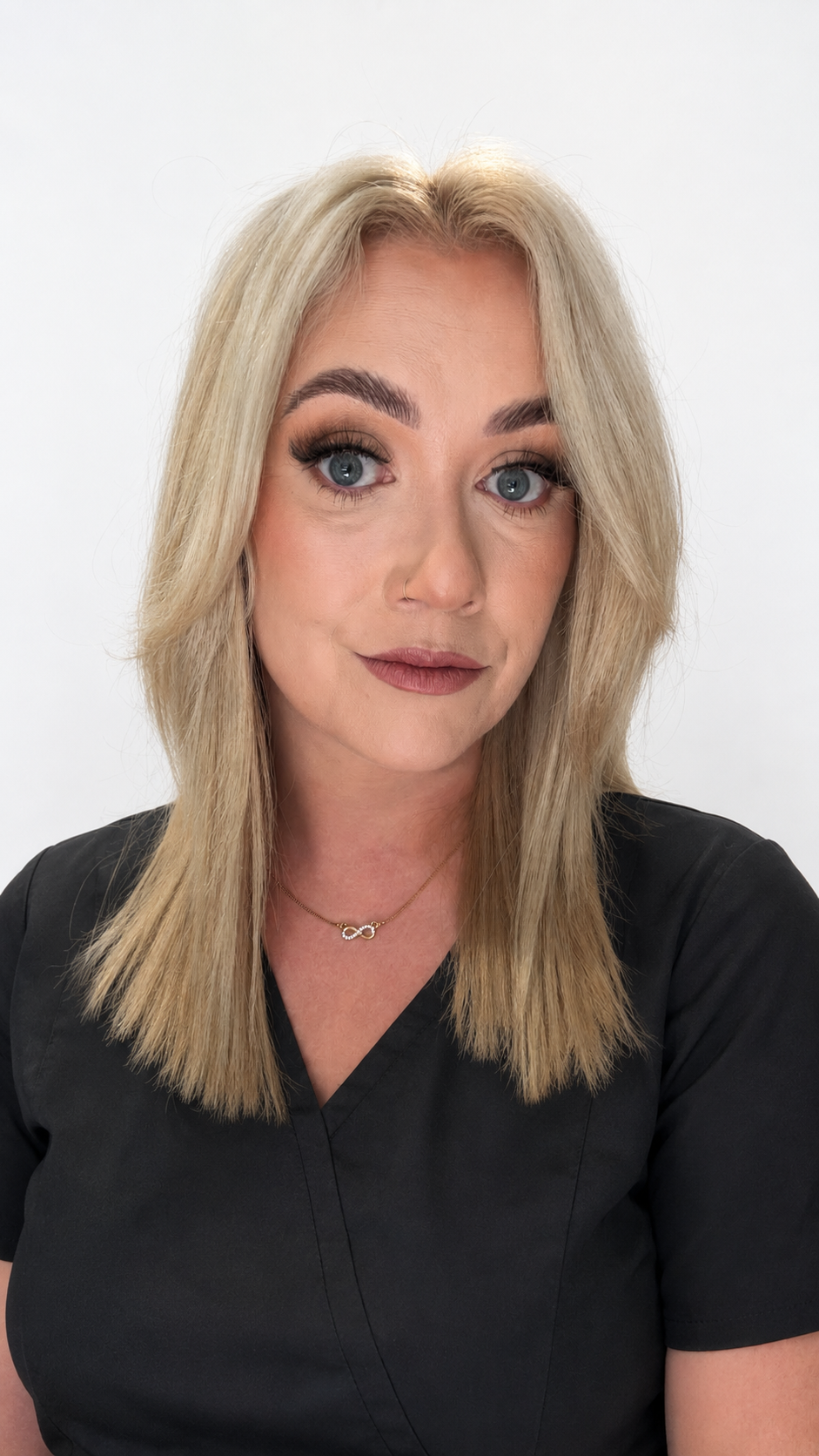 Close-up portrait of a woman with blonde hair and blue eyes, wearing a black top, a small nose hoop, and a gold infinity necklace, against a plain white background.