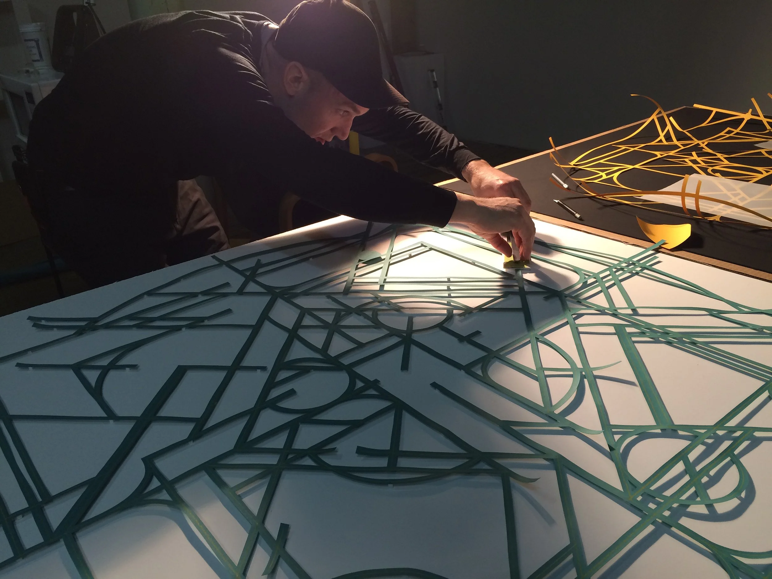 A man with a cap is working on a large illuminated geometric paper artwork with colorful, interconnected lines