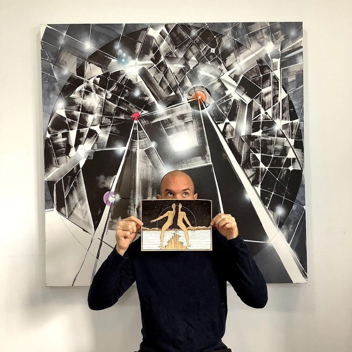Man holding a drawing in front of his face, with a large abstract black and white art piece on the wall behind him.