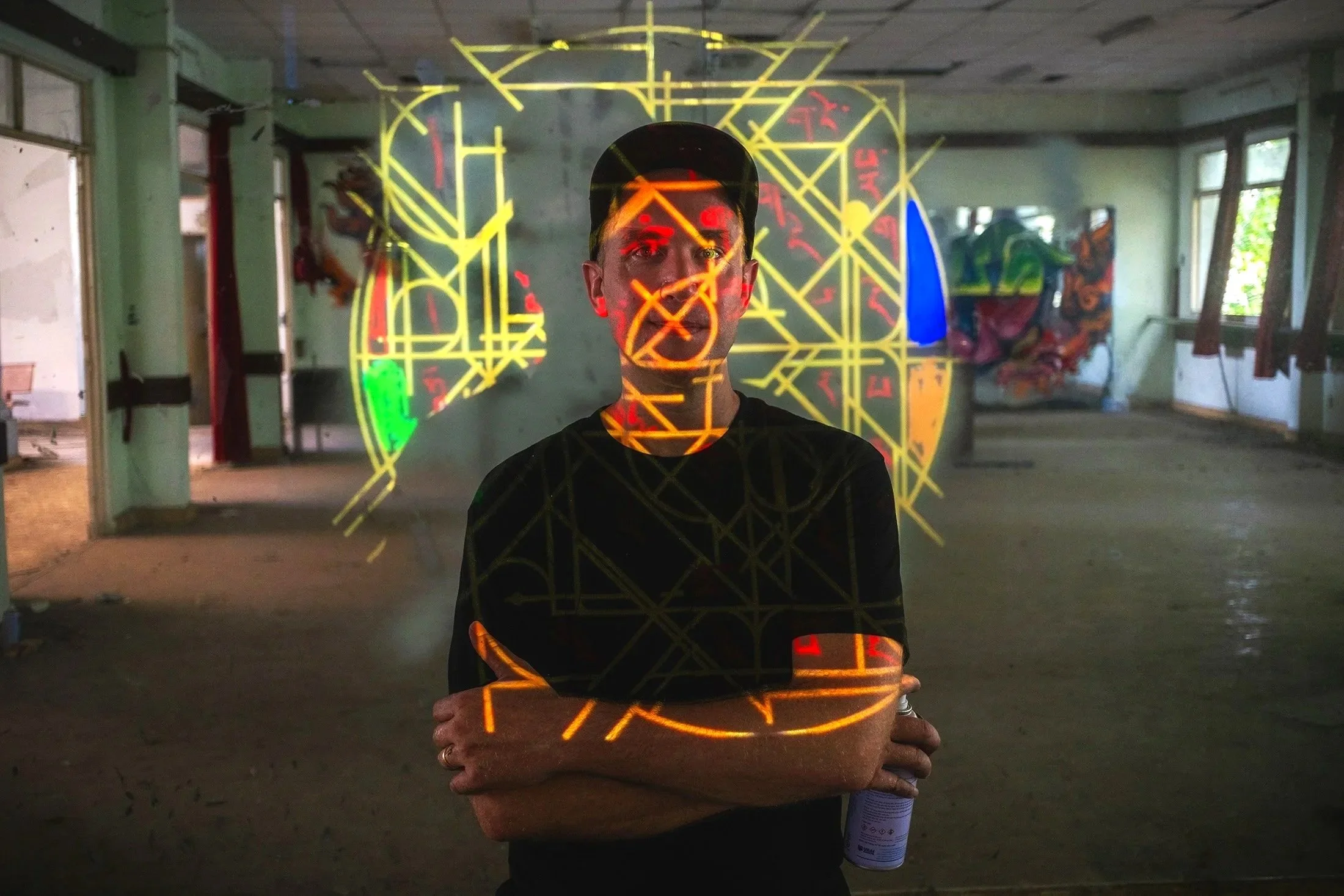 Person standing indoors with neon light projections on their face and body, arms crossed, in a spacious, unfinished room with windows and colorful graffiti on the walls.