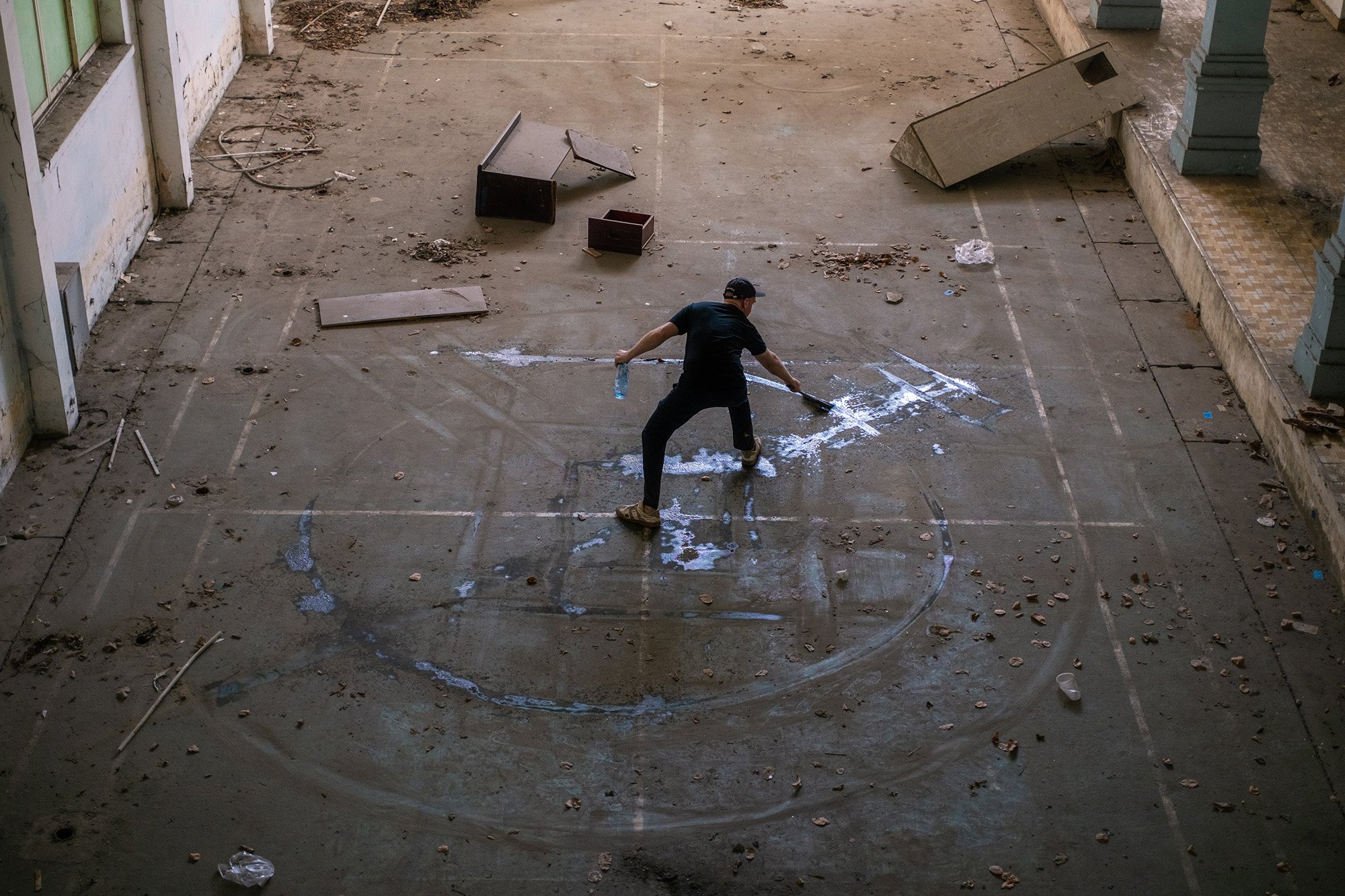 a man making an abstract geometric mandala artwork on a dirty floor using a broom and water