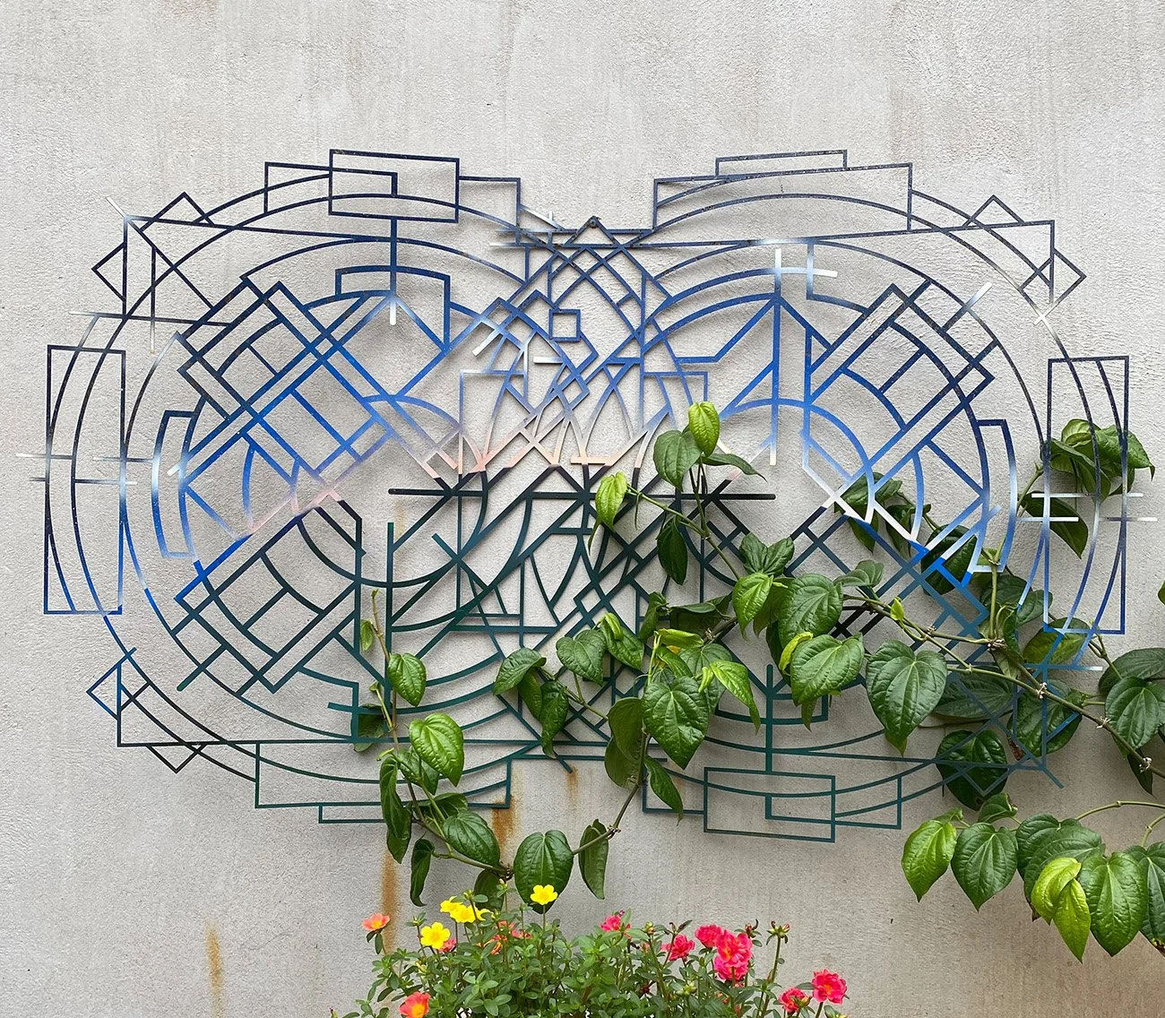 painted abstract geometric metal mandala sculpture mounted on an exterior wall with a plant growing across it