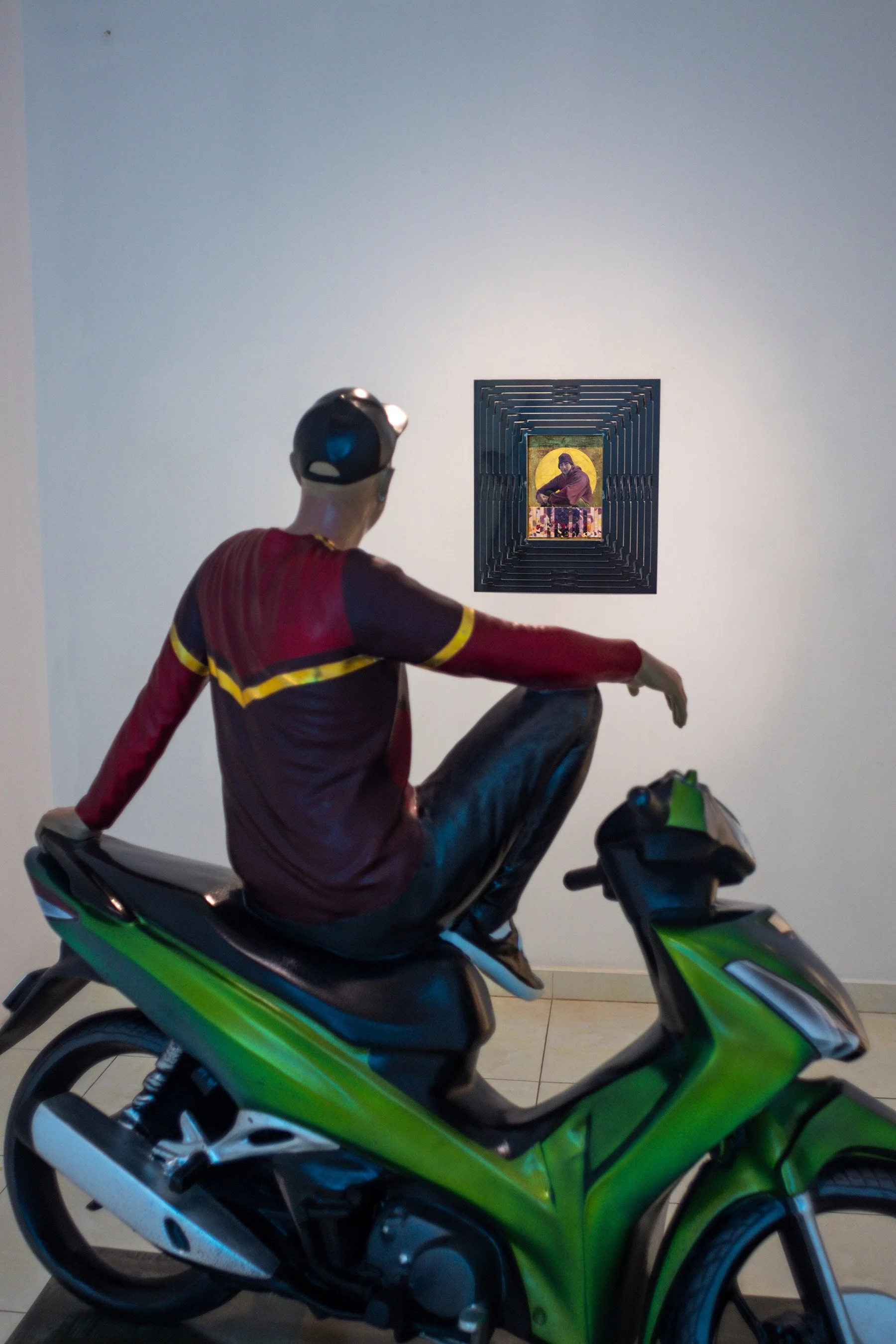 sculpture of a seated man on a motorbike looking at a painted portrait in the background