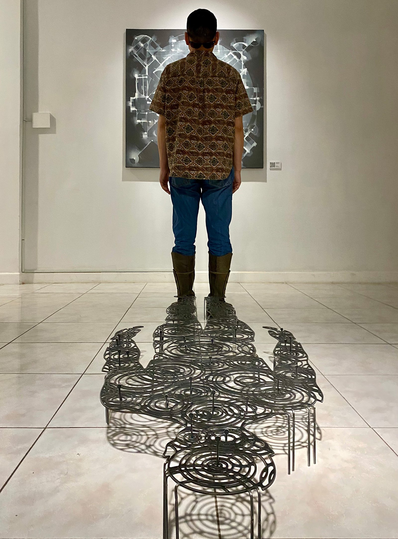 metal sculpture of a mans shadow made of rain drops on water in front of an abstract painting  with a viewer standing between the pieces in a gallery