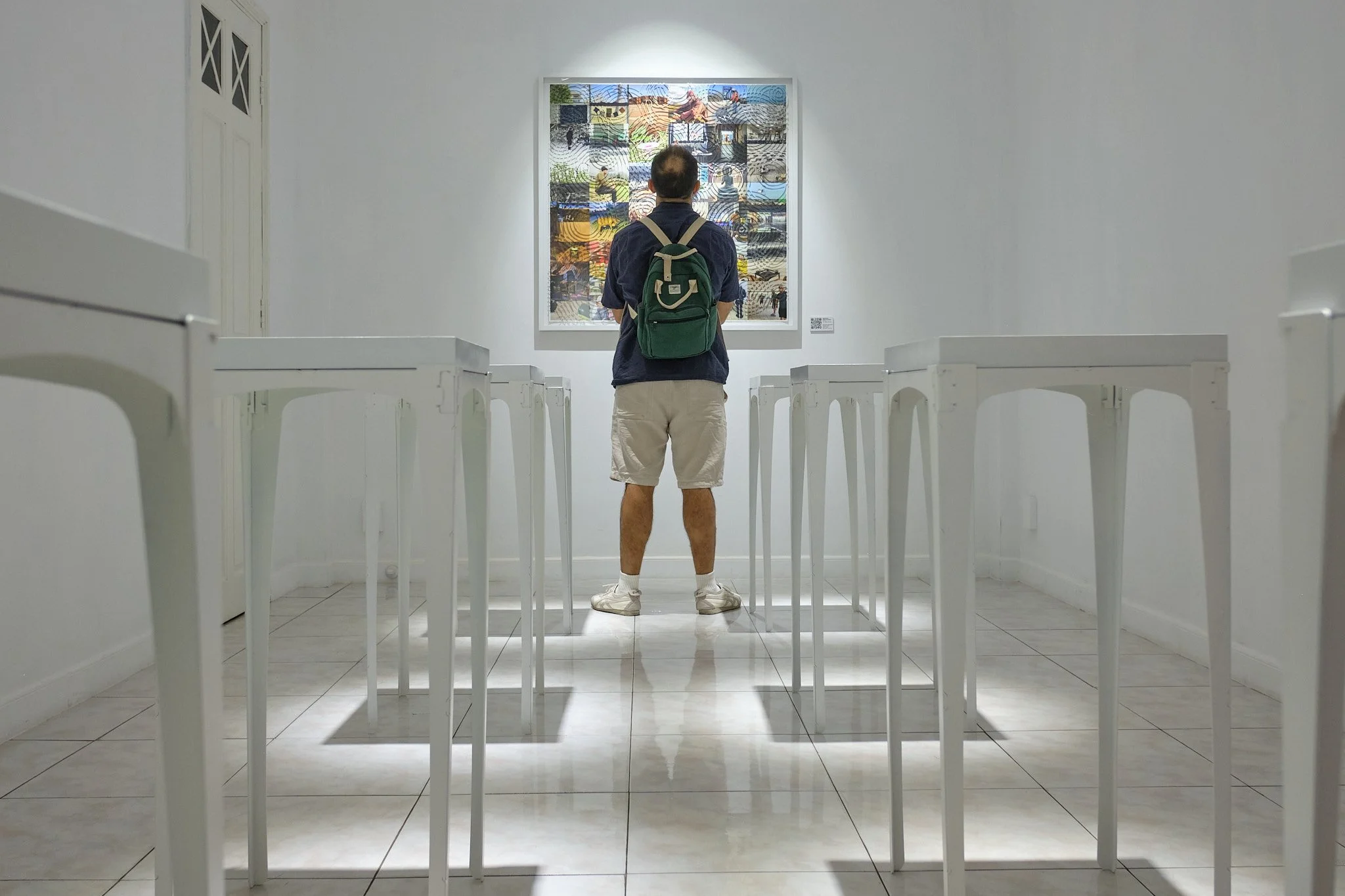man standing centered in a gallery installation between white tables looking at an artwork on the back wall