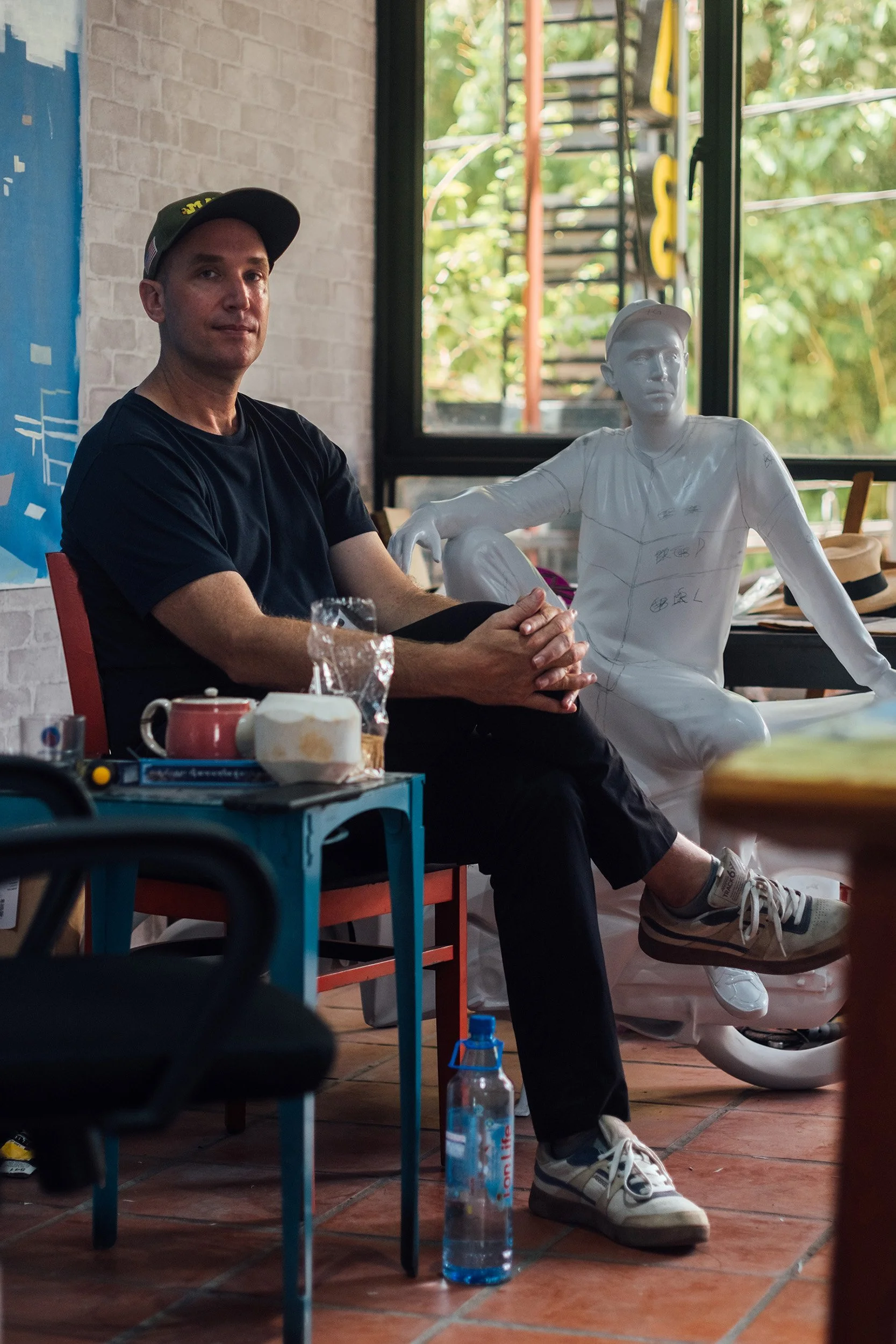 An artist sitting in his studio next to a white sculptural portrait of himself, with food, cups, and personal items on a table nearby, and a bottle of water on the floor, with a window showing greenery outside.
