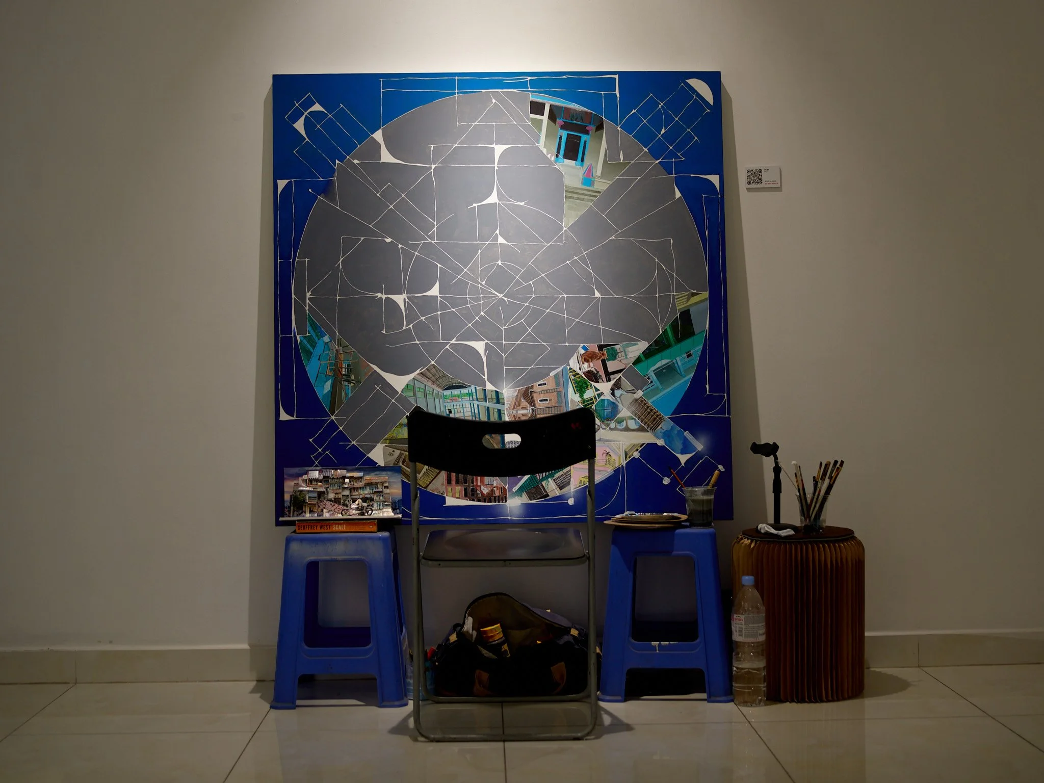 Art exhibit with a large mirror mosaic on blue background, partially reflecting a building and street scene, flanked by blue chairs, with various objects including a water bottle, bag, and a table with art supplies.