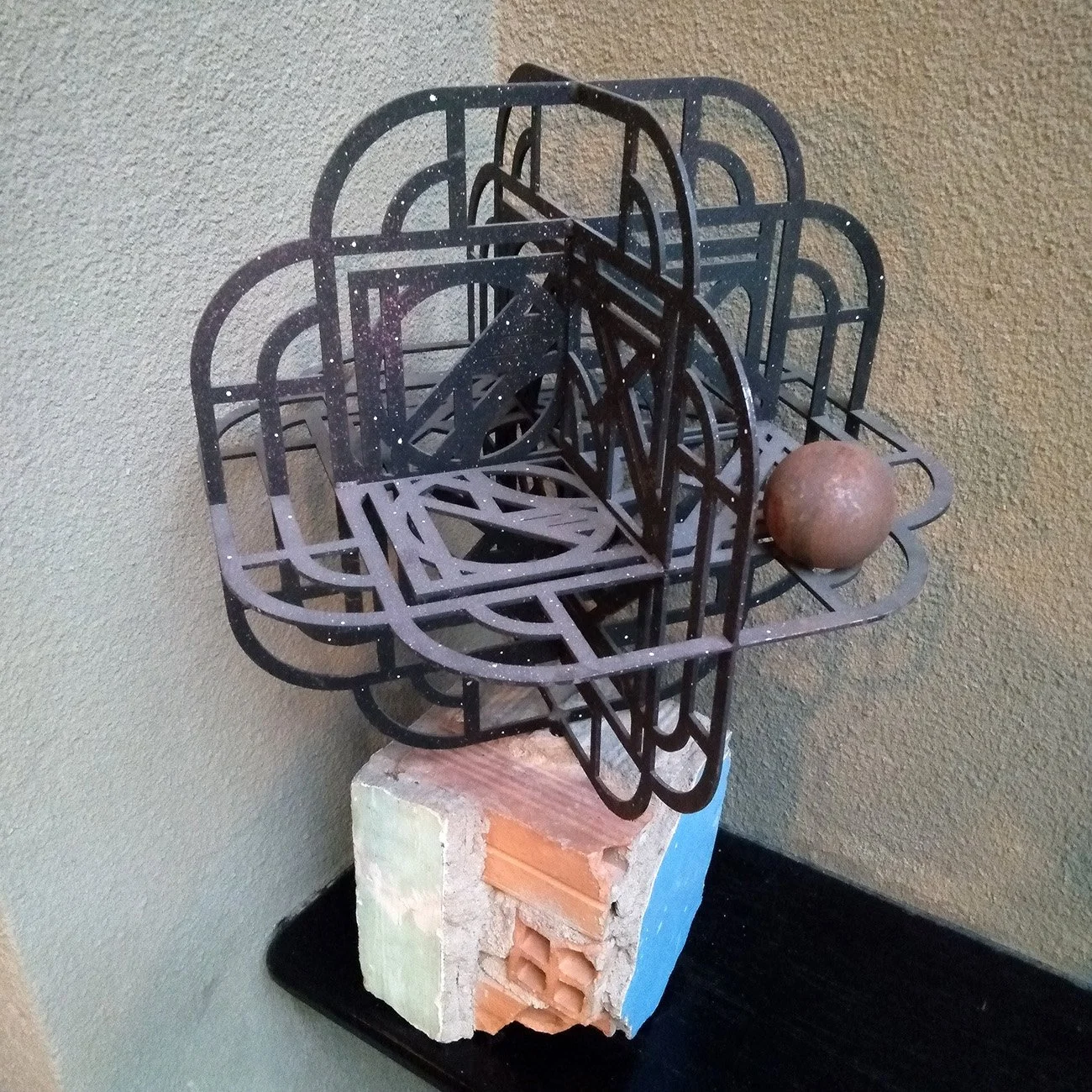 abstract geometric metal sculpture painted black mounted on a block of painted bricks with a metal sphere resting on top