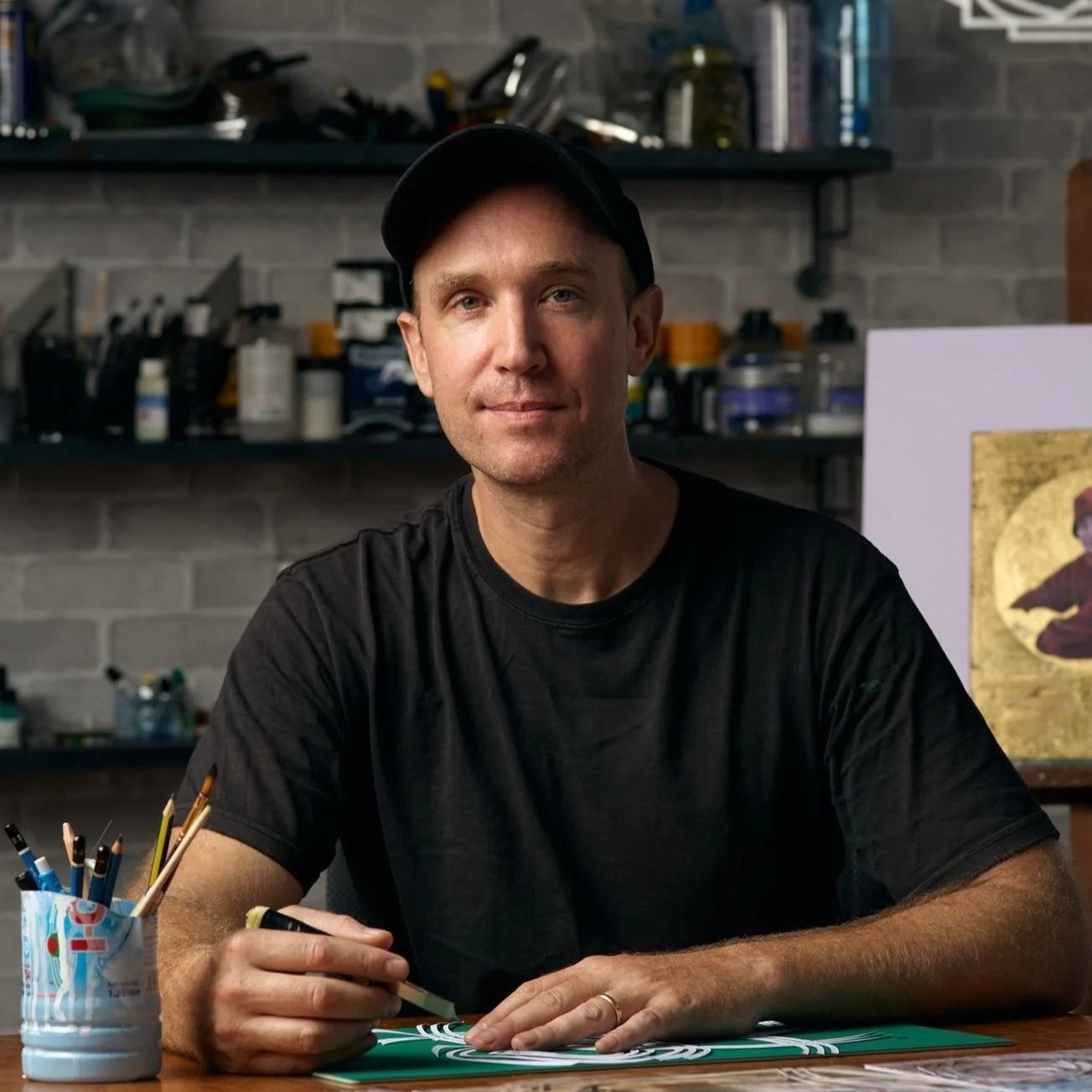 A man sitting at a table in a workshop, holding a matte knife and working on an art project with a focused expression.