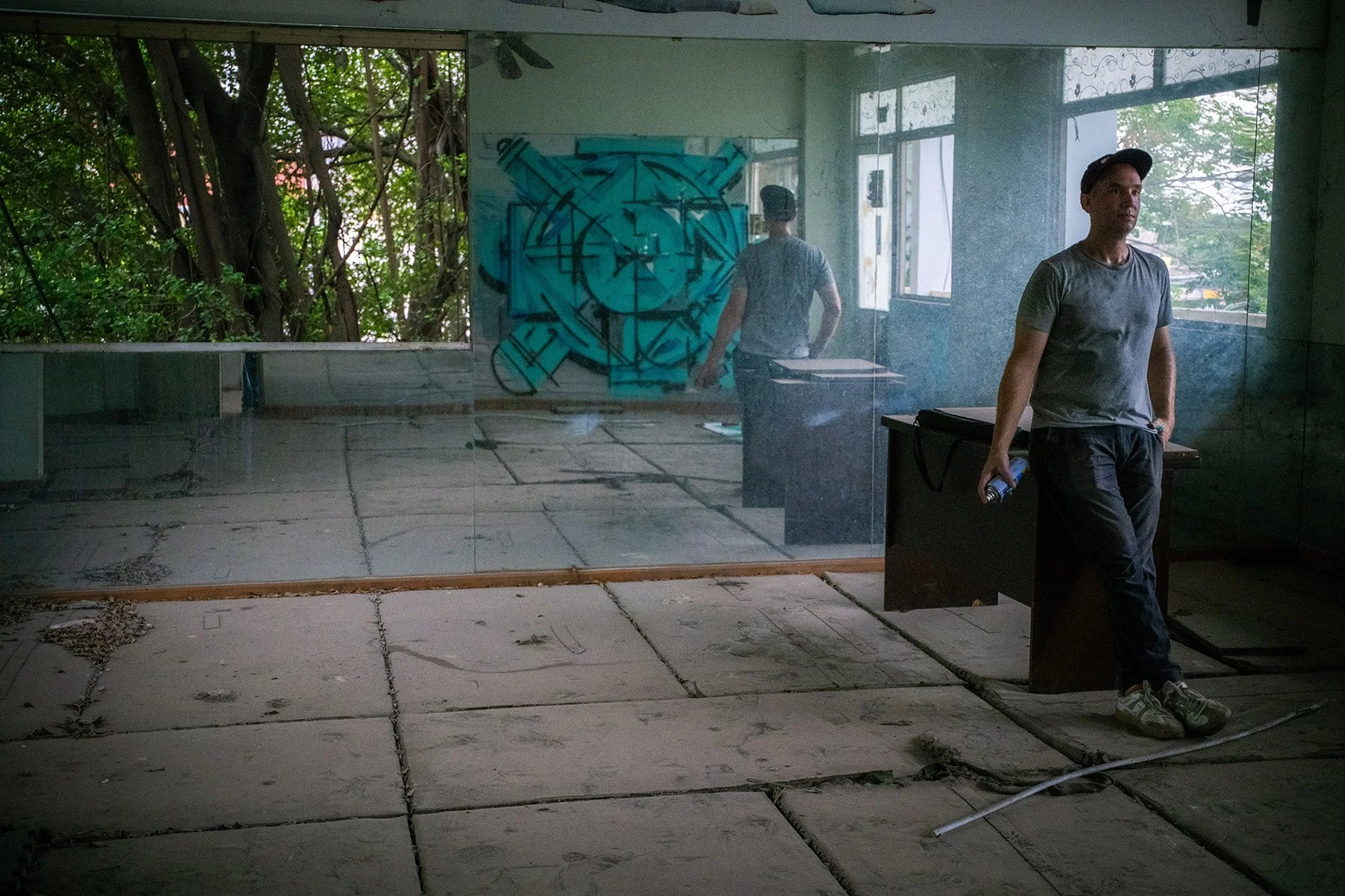 man holding spray paint resting looking at an abstract geometric mandala on a mirror in the room of an abandoned building