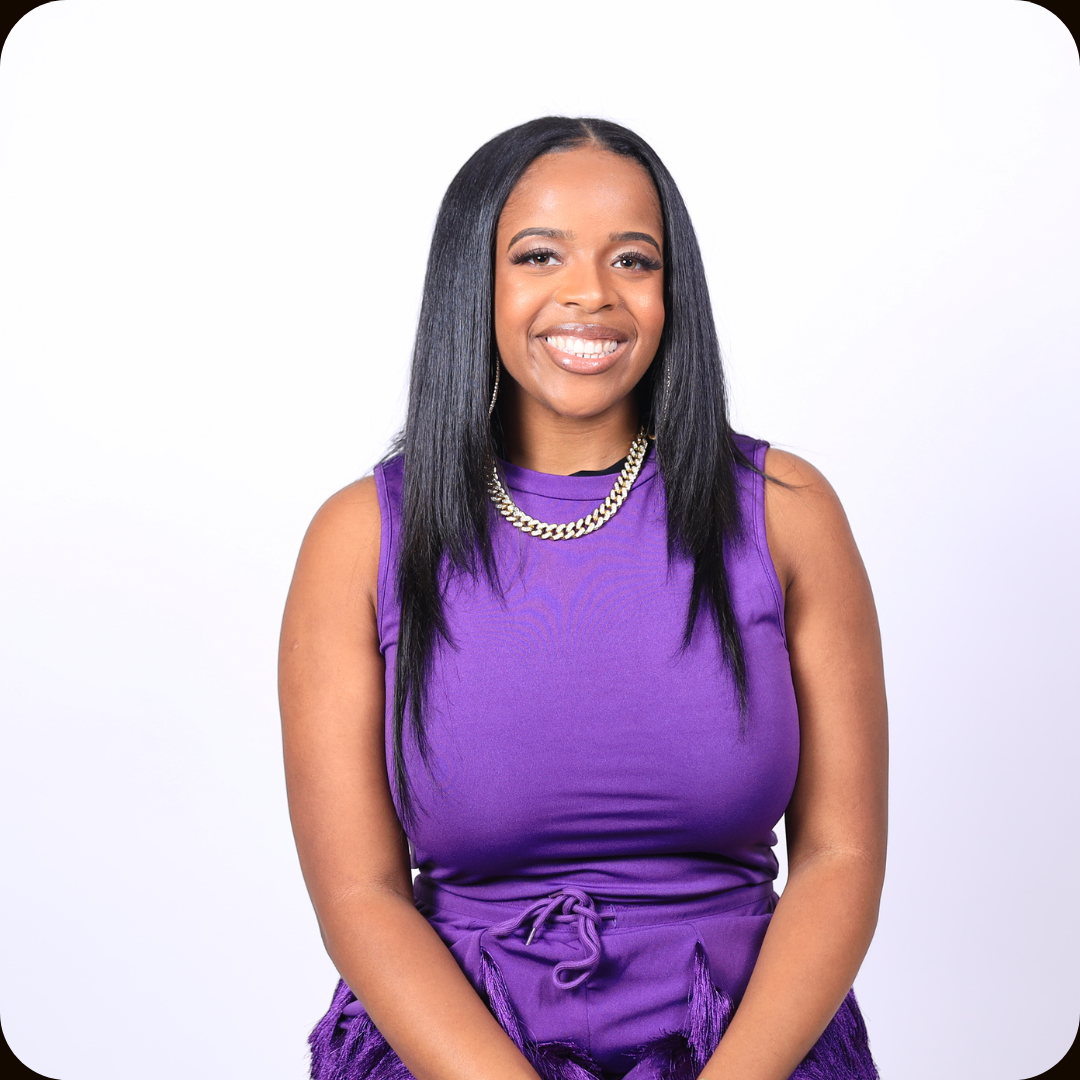 A woman with long black hair and medium skin tone smiling, wearing a purple sleeveless dress, a double gold chain necklace, and earrings, standing against a white background.