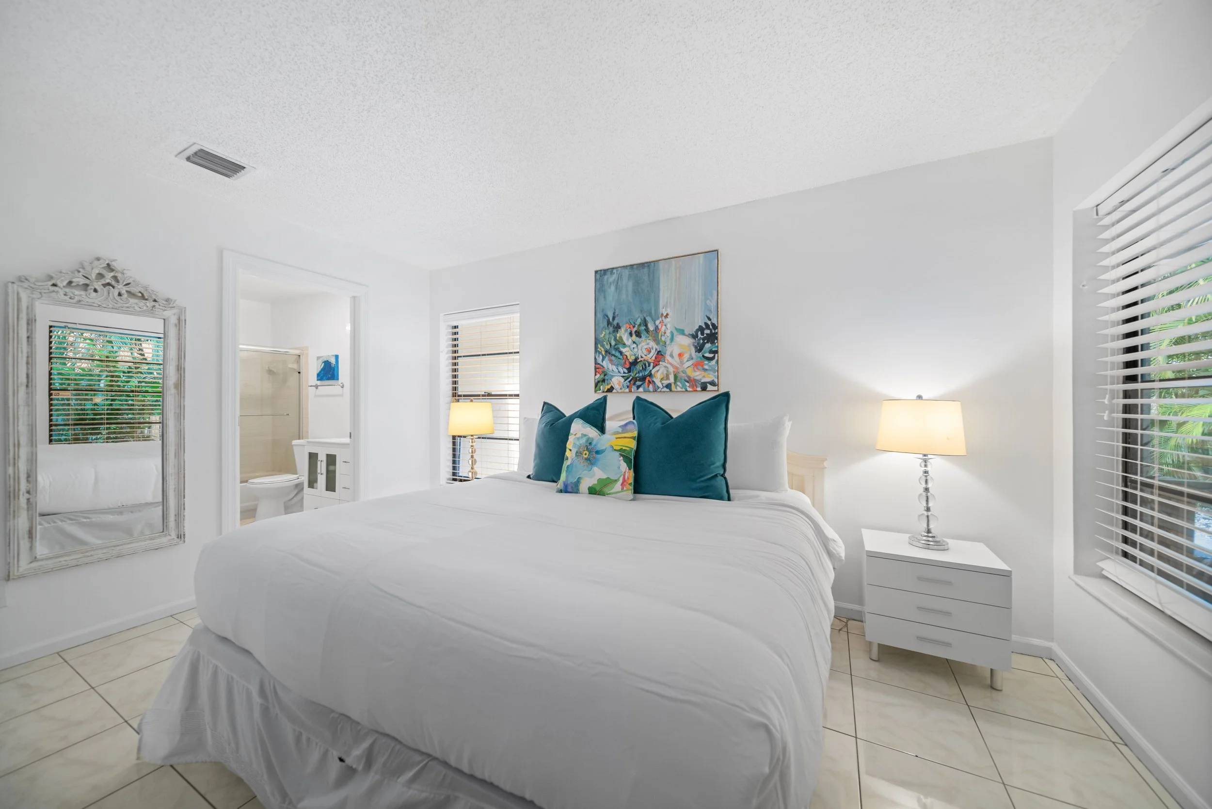 Bright white bedroom with modern decor, featuring a bed with colorful pillows, two lamps, artwork on the walls, and an ensuite bathroom visible through an open door.