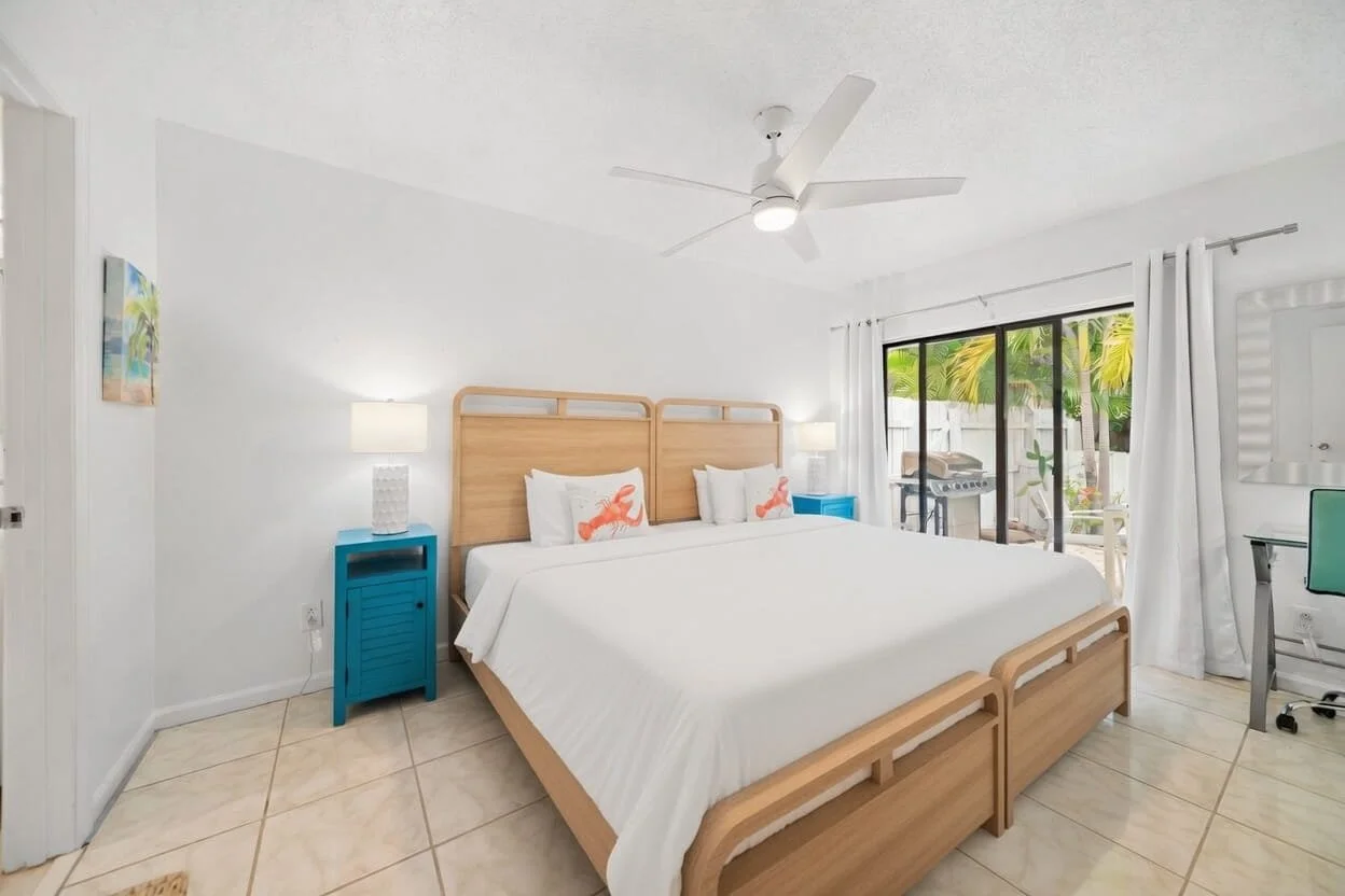 A bright hotel room with a large bed, two blue nightstands with white lamps, and a sliding glass door leading to an outdoor patio with yellow and green plants.