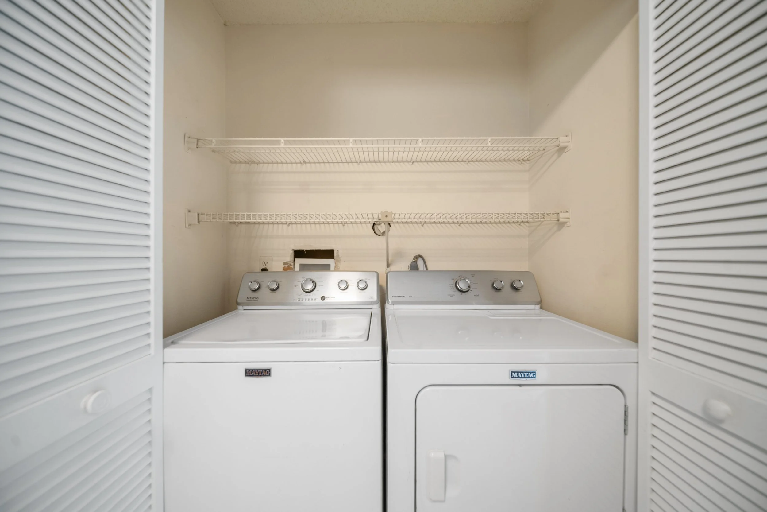 A laundry closet with closed louvered doors, featuring a washer and dryer beneath two empty wire shelves.