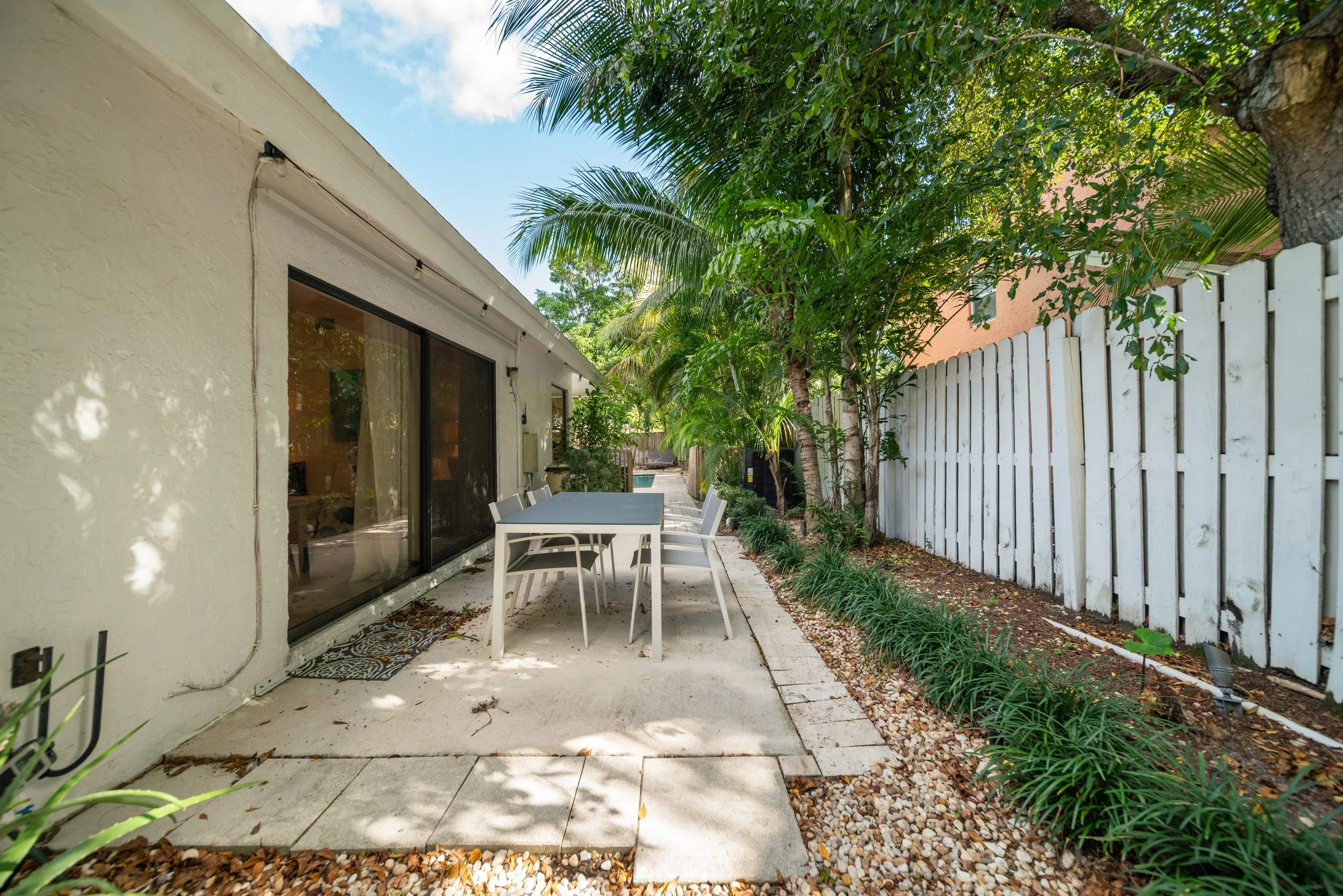 A backyard patio area featuring a white table with chairs, surrounded by lush green trees and plants, with a white fence and a house with large sliding glass doors in the background.