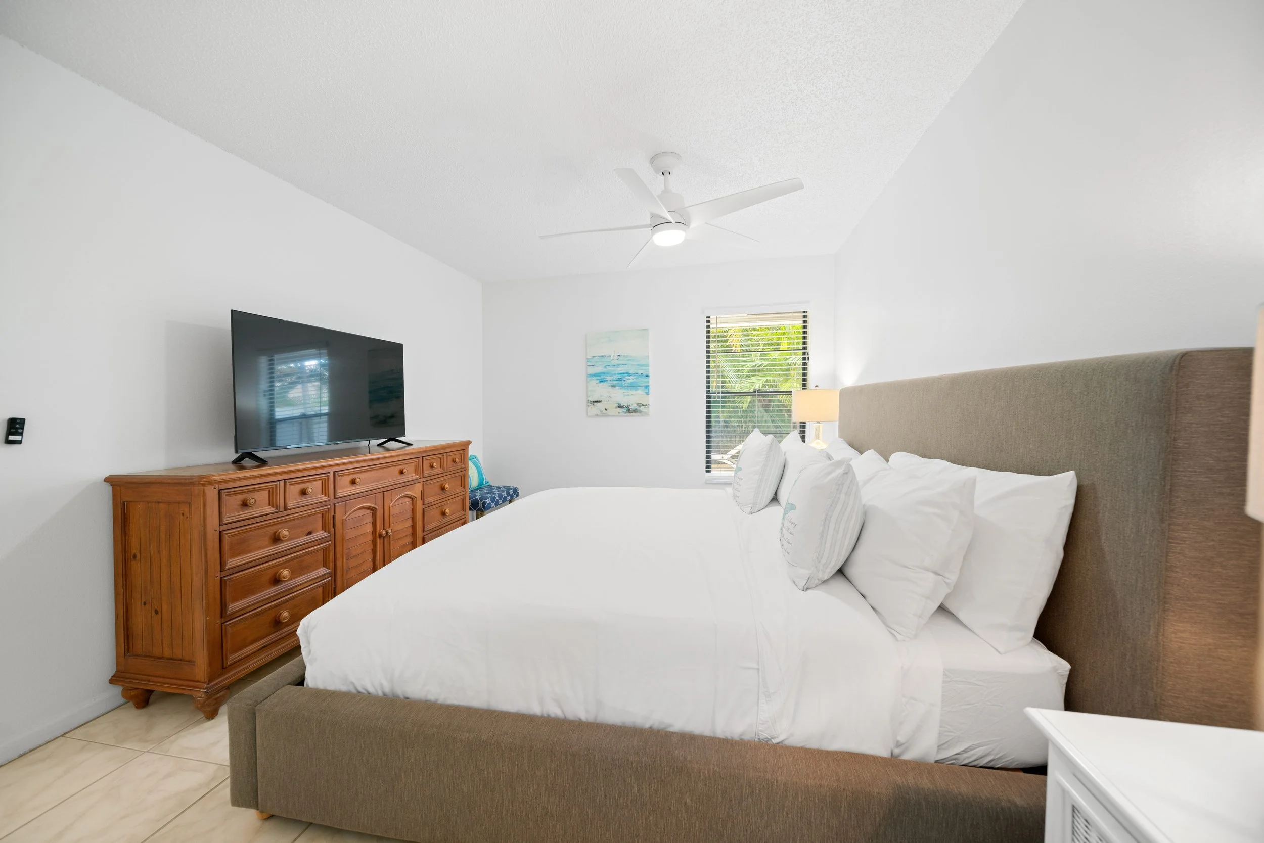 A bedroom with a large bed, beige upholstered headboard, white bedding, and pillows. There is a wooden dresser with a TV on top, a small blue patterned bench, a window with blinds, and a ceiling fan with a light. A piece of art hangs on the wall.