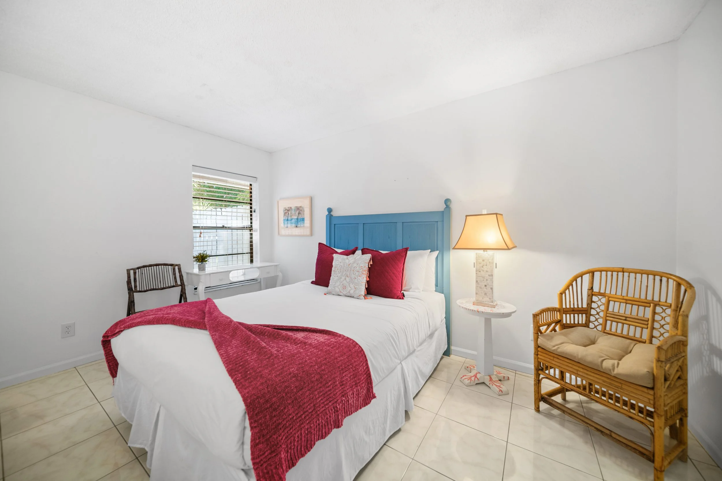 A bedroom with white walls and tiled floor. There is a bed with white bedding, purple and white pillows, and a red throw blanket. A blue headboard is behind the bed. Next to the bed is a white round side table with a lamp, and a wicker chair with a c