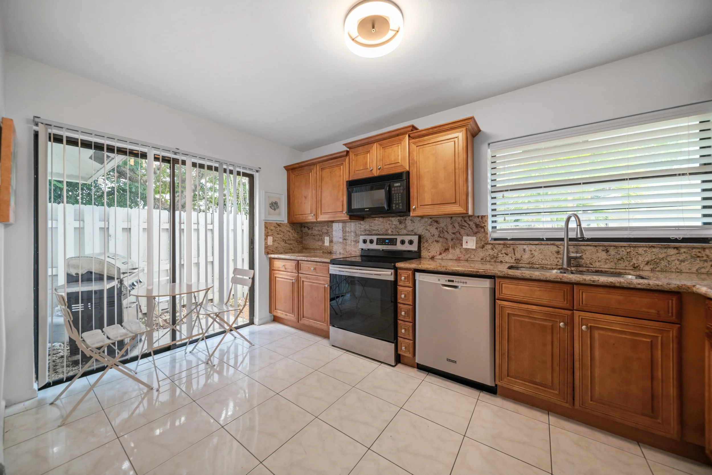 Kitchen with wooden cabinets, a black stove, microwave, dishwasher, granite countertops, window blinds, tiled floor, and a sliding glass door leading to a patio with a barbecue grill.