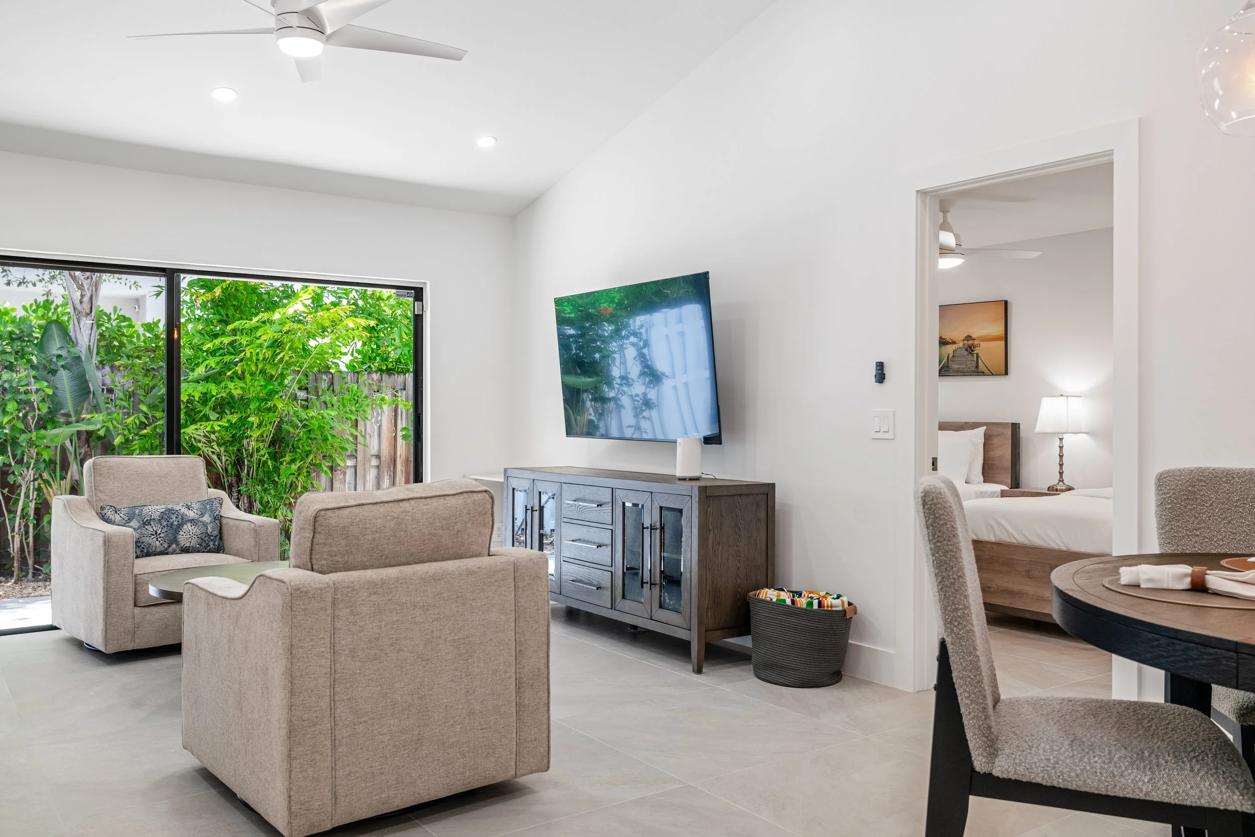 Living room with beige sofas, a wooden gray TV console with glass doors, a wall-mounted flat-screen TV, a sliding glass door with greenery outside, and a sitting area in a bedroom with a bed, a bedside lamp, and a framed ocean sunset picture.