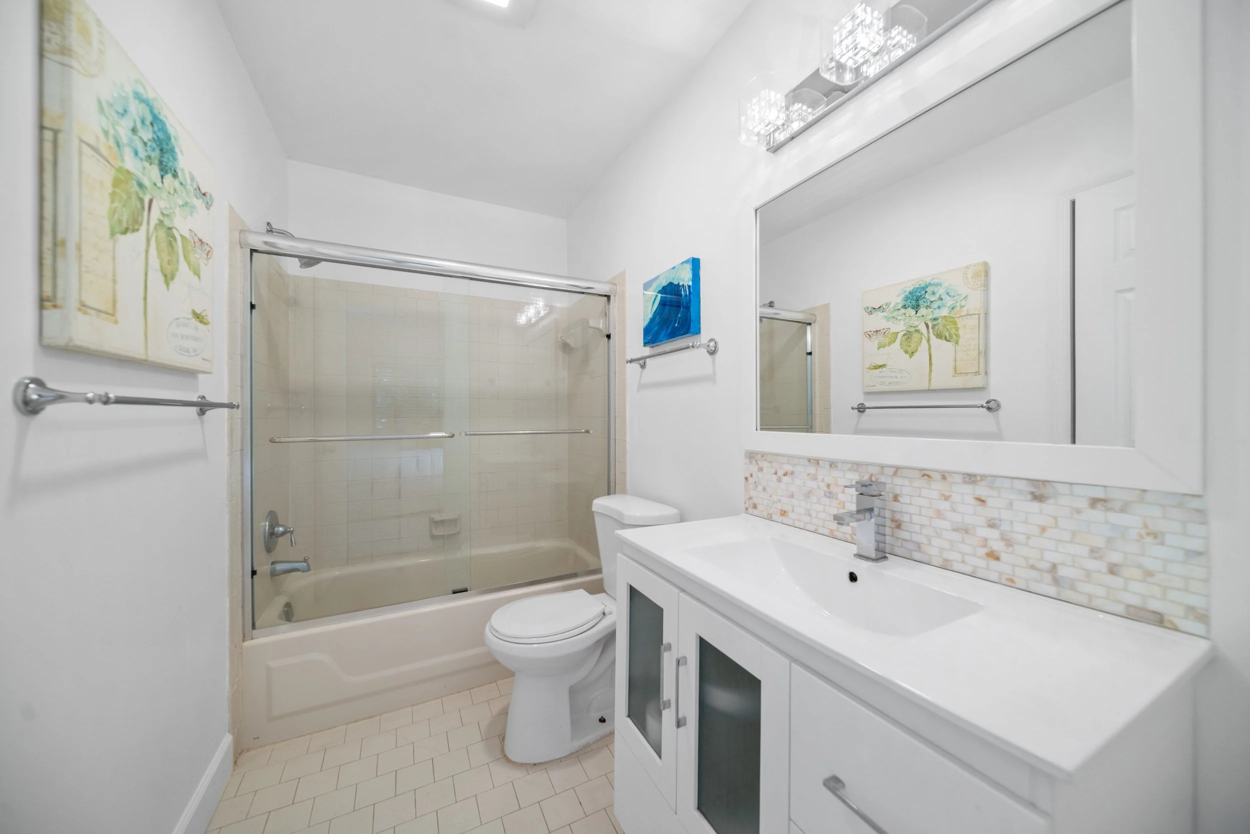 A small bathroom with a beige bathtub and sliding glass door, toilet, white vanity with a large mirror, two decorative paintings, and a tiled floor, all in a modern style.