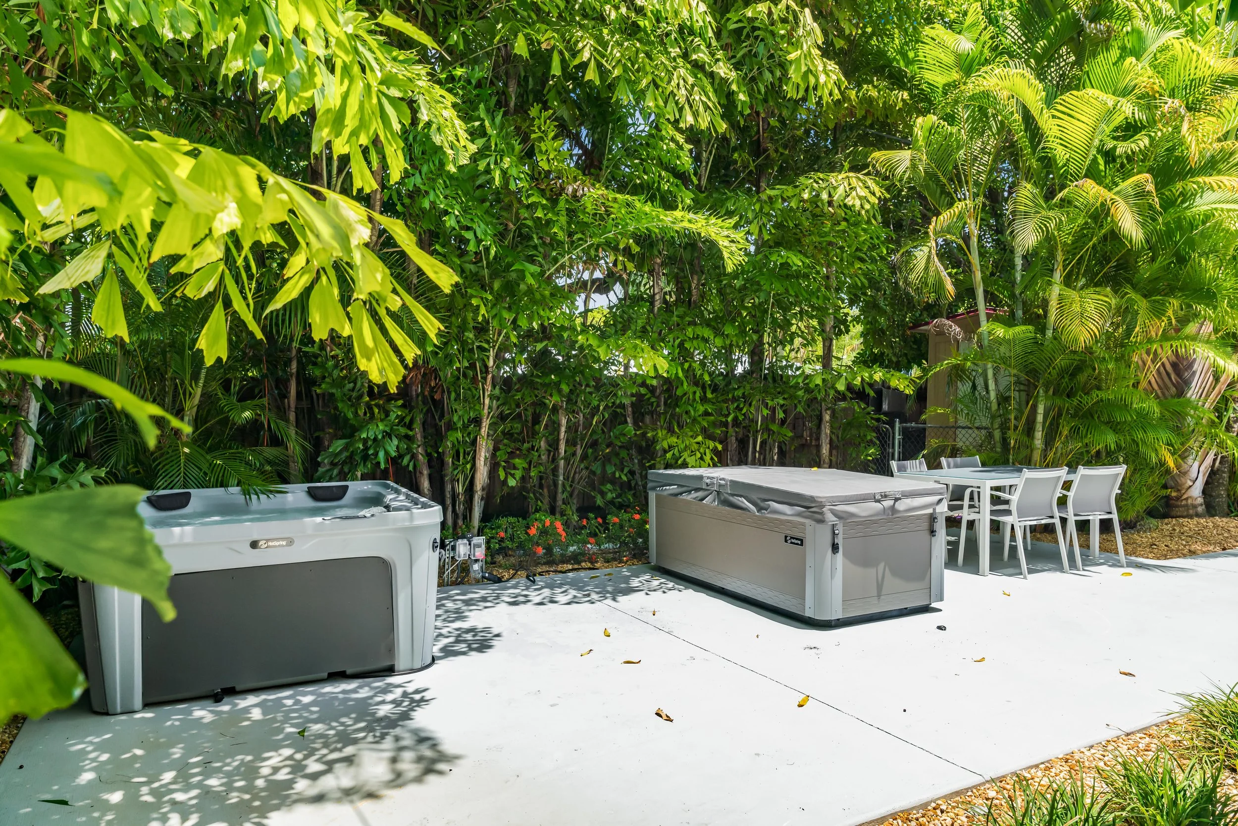 Backyard patio with outdoor furniture, large hot tubs or storage containers, and tall lush trees and plants providing shade.